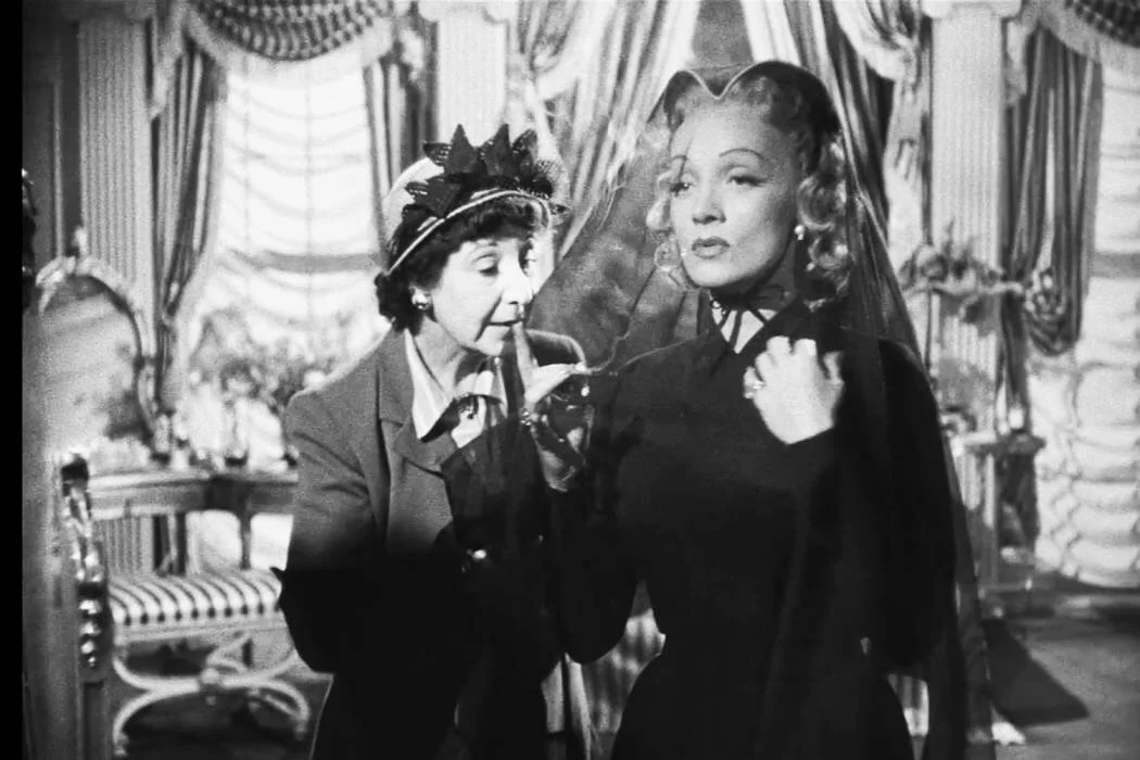 Marlene Dietrich’s character Charlotte Inwood is wearing an elegant black fitted dress with sharp black pumps. This is accessorized with a black hat and long dramatic veil. 
