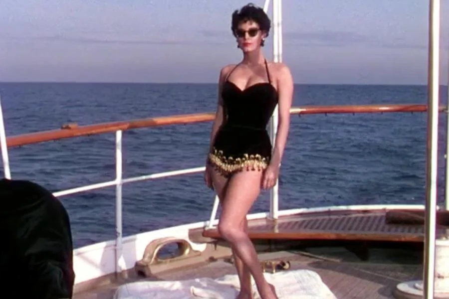 Ava Gardner’s character Maria wears a halter-top style one piece bathing suit with gold fringe detailing around the legs as she suns herself on the yacht deck.
