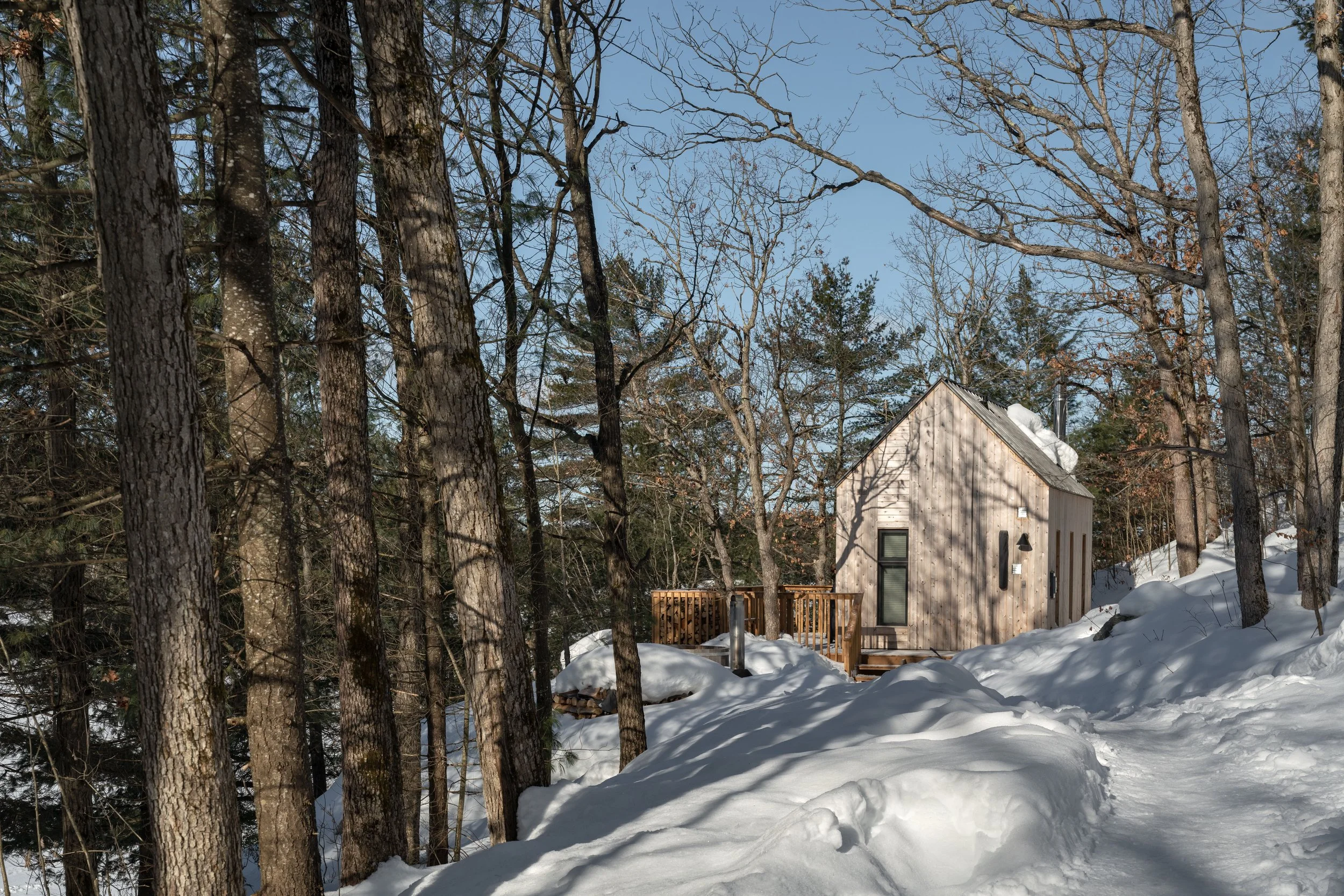 A small wooden cabin with a snow-covered roof in a snowy, forested area with tall trees and a clear blue sky.