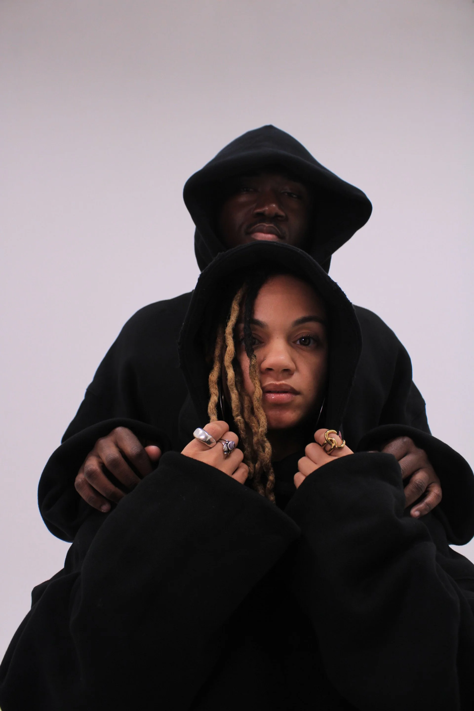 Couple in black oversized VOUFO hoodies with hoods up, he stands behind her, against a lilac backdrop