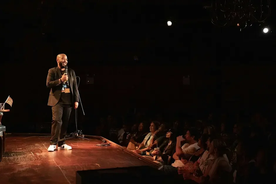 A comedian stands on stage holding a microphone, performing in front of a seated audience in a dimly lit venue.