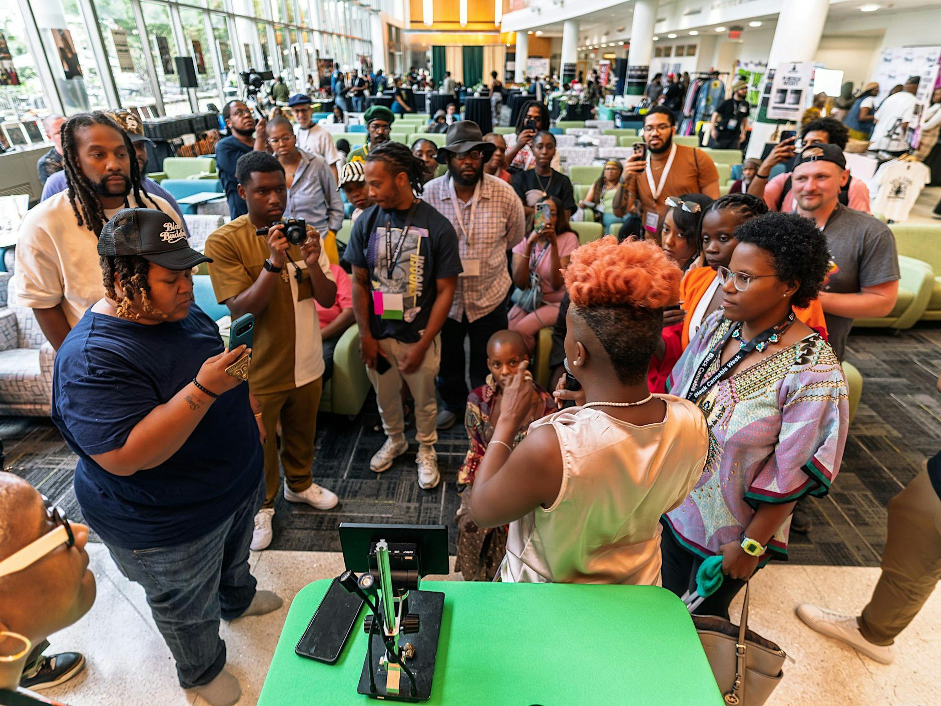A diverse group of people gathered around a table at an indoor event or conference. Some are taking photos or videos, while others are watching or listening. The scene is lively with booths and displays in the background.