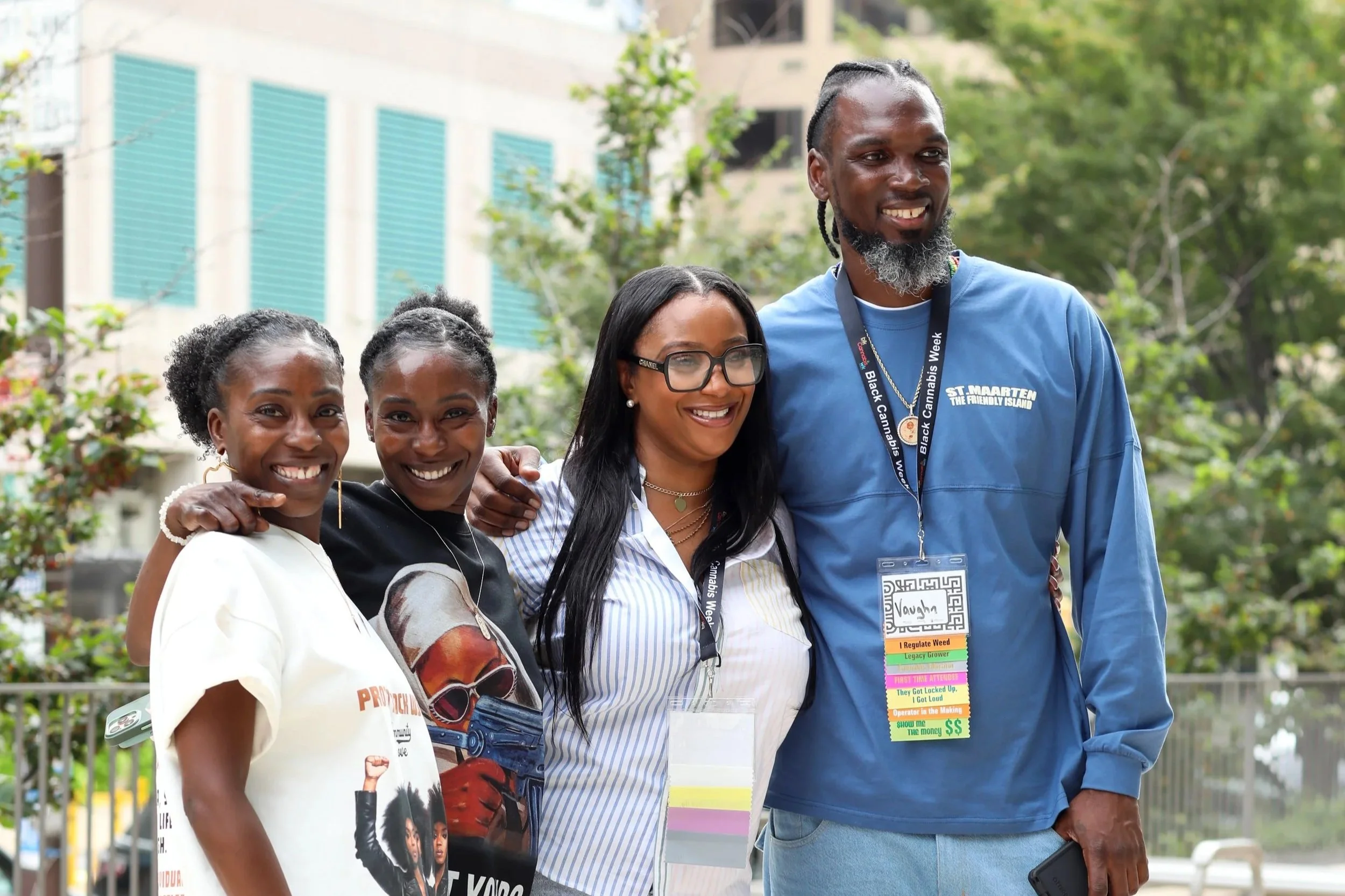 Group of four diverse people outdoors smiling, with trees and buildings in the background.