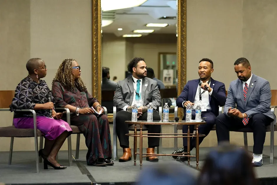Five individuals seated on a stage, participating in a panel discussion with water bottles in front of them, in a conference setting.