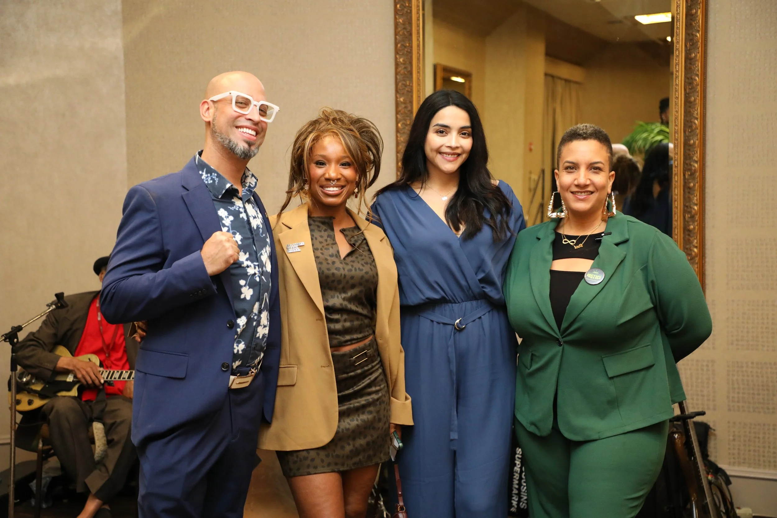Four diverse women smiling and standing together in formal attire at an event, with a musician playing guitar in the background and a large mirror on the wall behind.