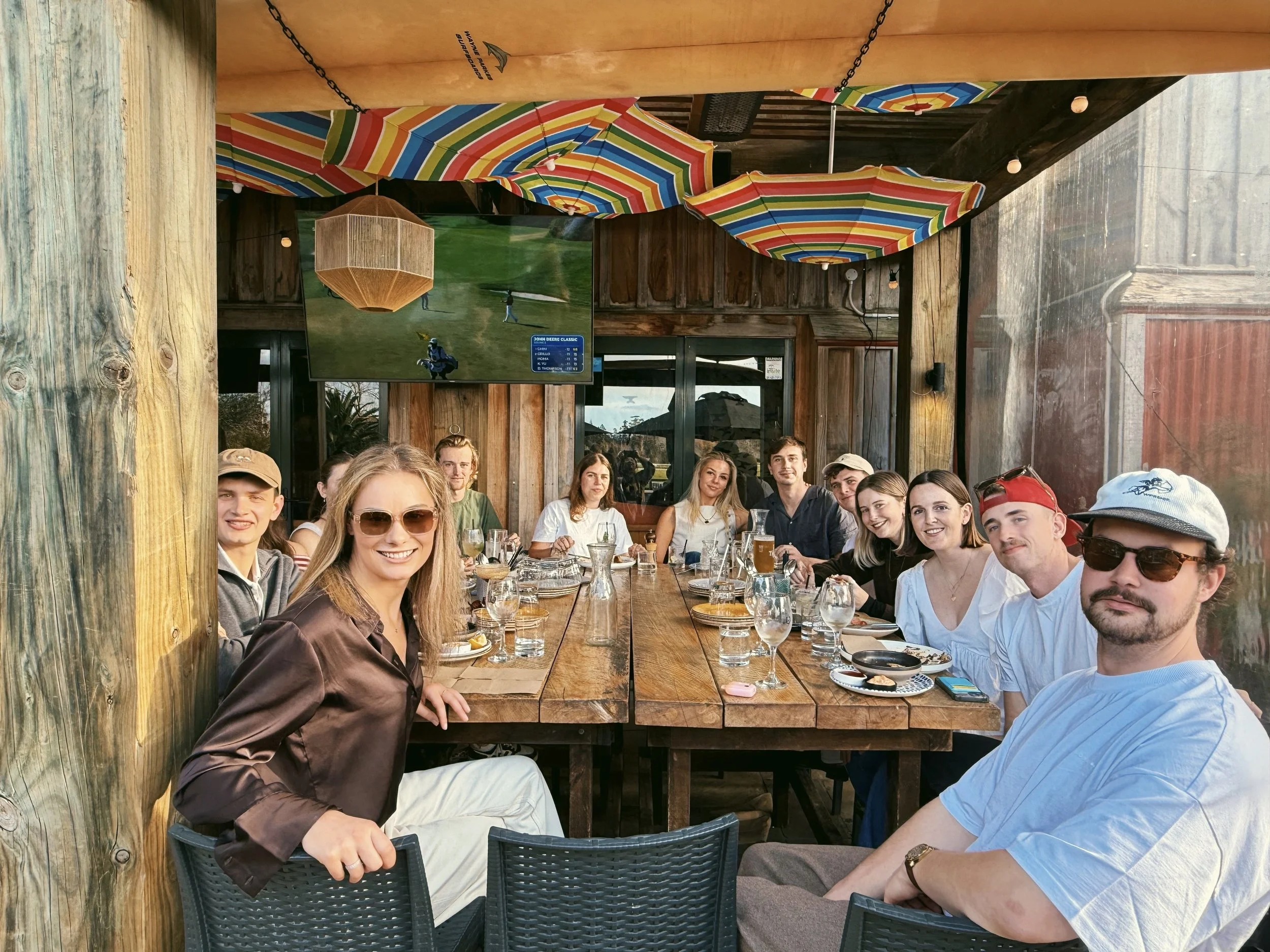 Group of ten people sitting at a long wooden table outdoors, enjoying drinks and food, with colorful umbrellas overhead and a large television screen on the wall.