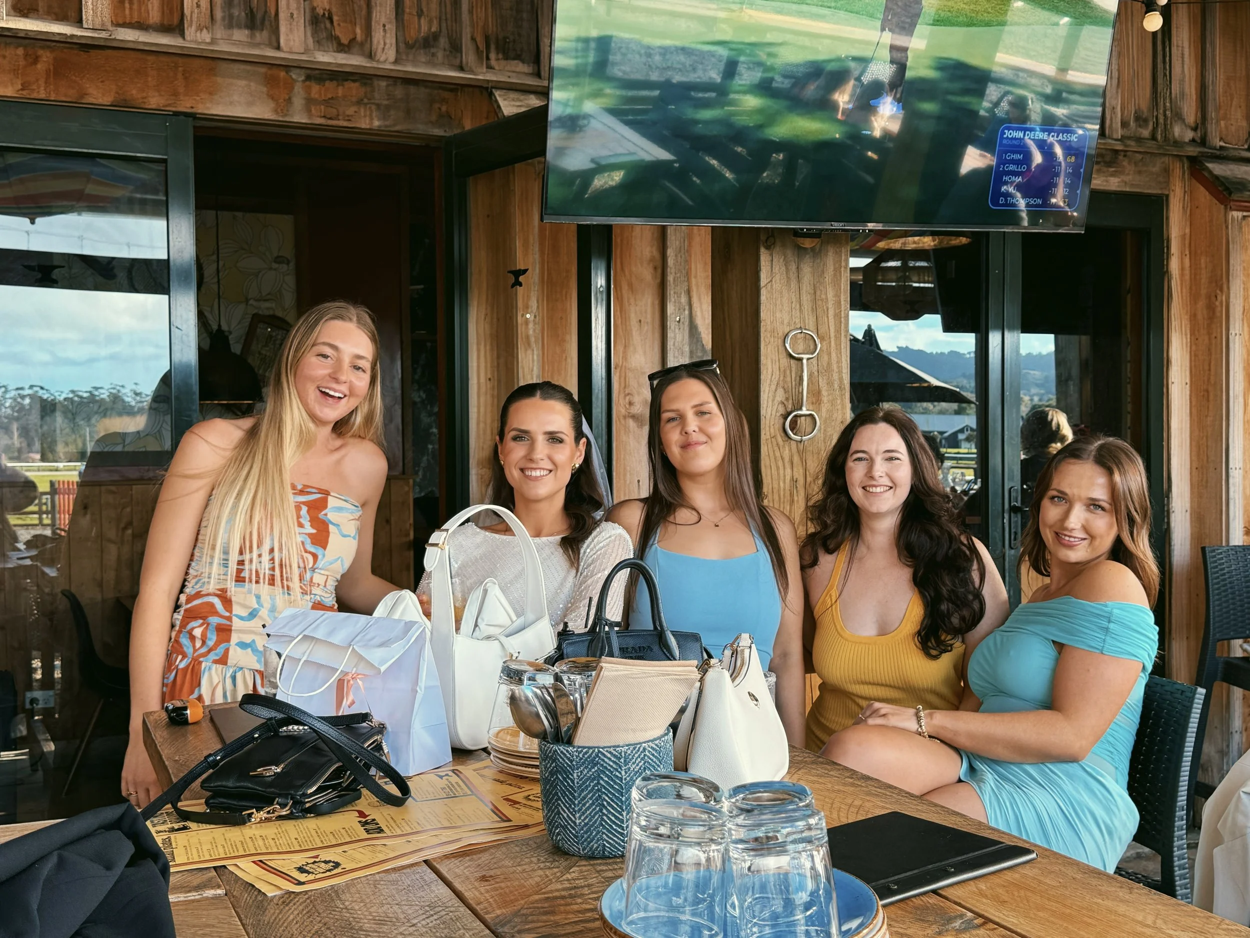 Five women sitting at a restaurant table, smiling and posing for the photo. The table has various bags, glasses, and menus. The restaurant has wooden walls and large windows showing outdoor scenery. A TV screen is mounted on the wall above the women.