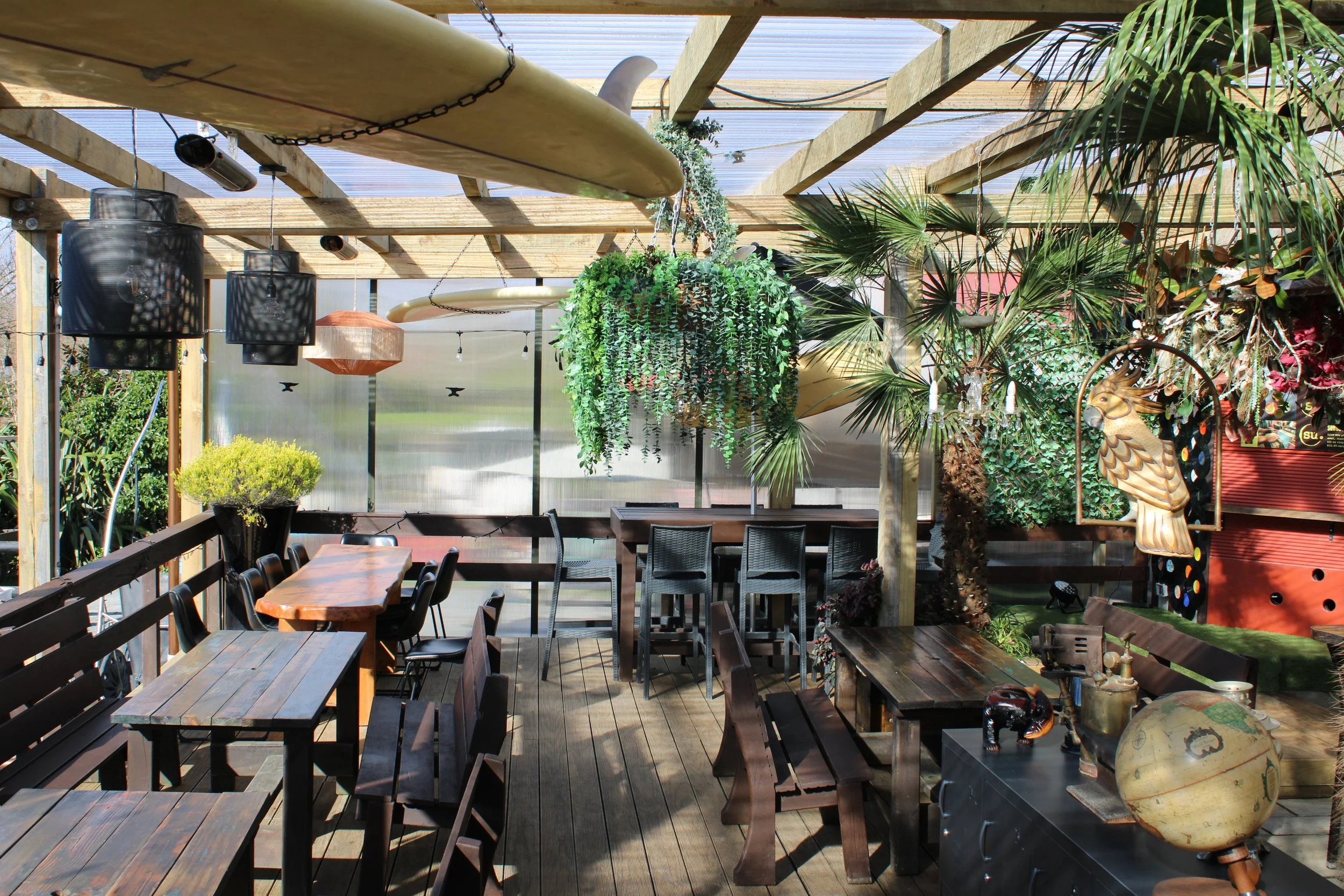 Outdoors cafe with wooden tables, benches, hanging plants, decorative surfboards, and a tropical theme.
