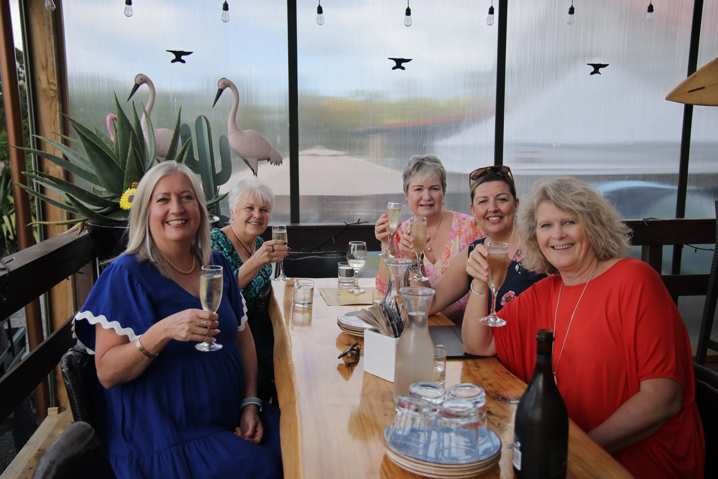 Five women celebrating with drinks at a long wooden table inside a sunroom with decorative flamingos and cacti on the window ledge.