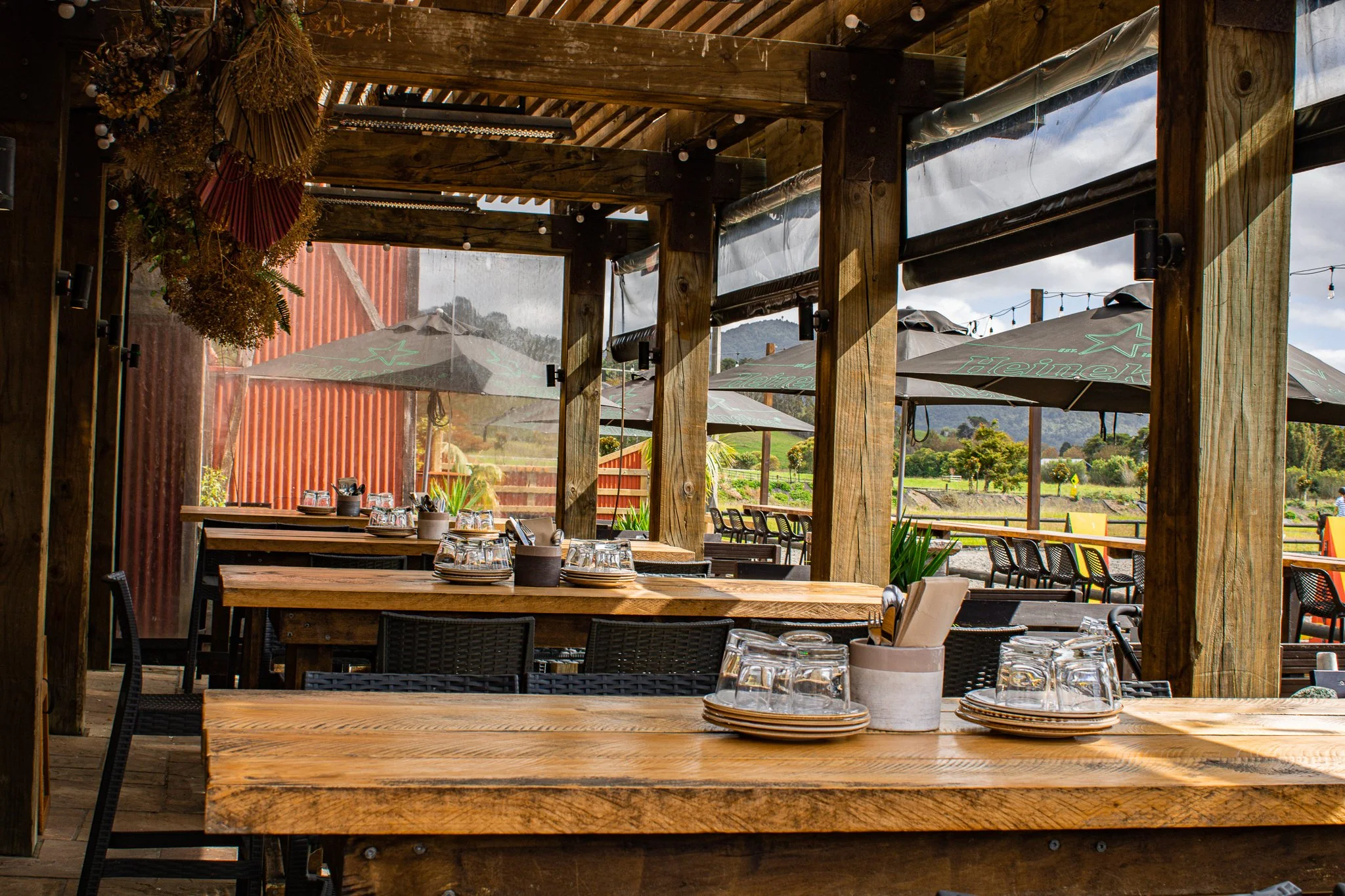 Outdoor restaurant seating area with wooden tables, chairs, and glassware. Overhead wooden beams provide shade, and the view includes a rustic red building and green outdoor umbrellas.