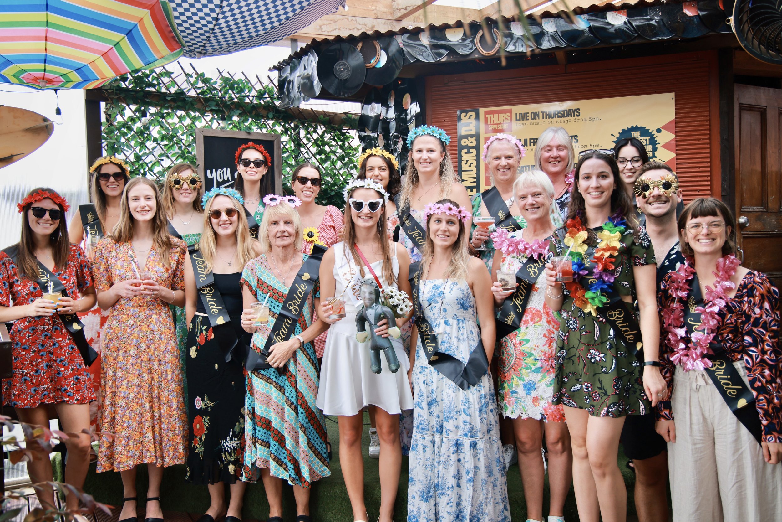 Group of women wearing floral dresses and flower crowns, celebrating on a decorated outdoor patio with drinks, some with sashes and leis, smiling for a photo.