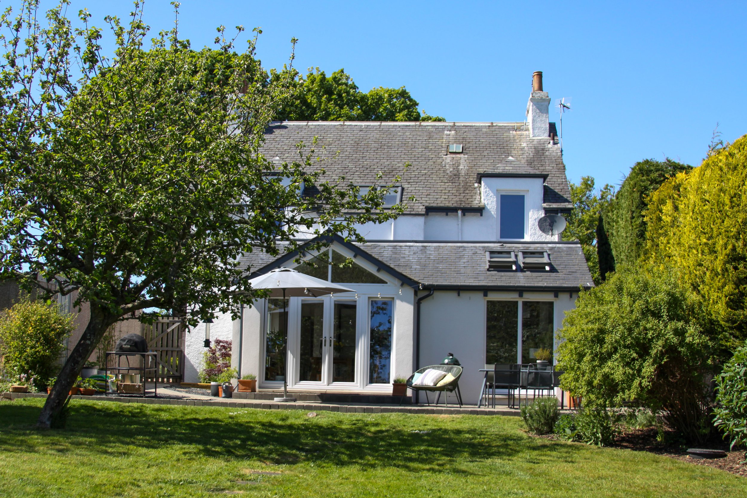 A white house with a gray roof and a garden with trees, bushes, and a patio with outdoor furniture.