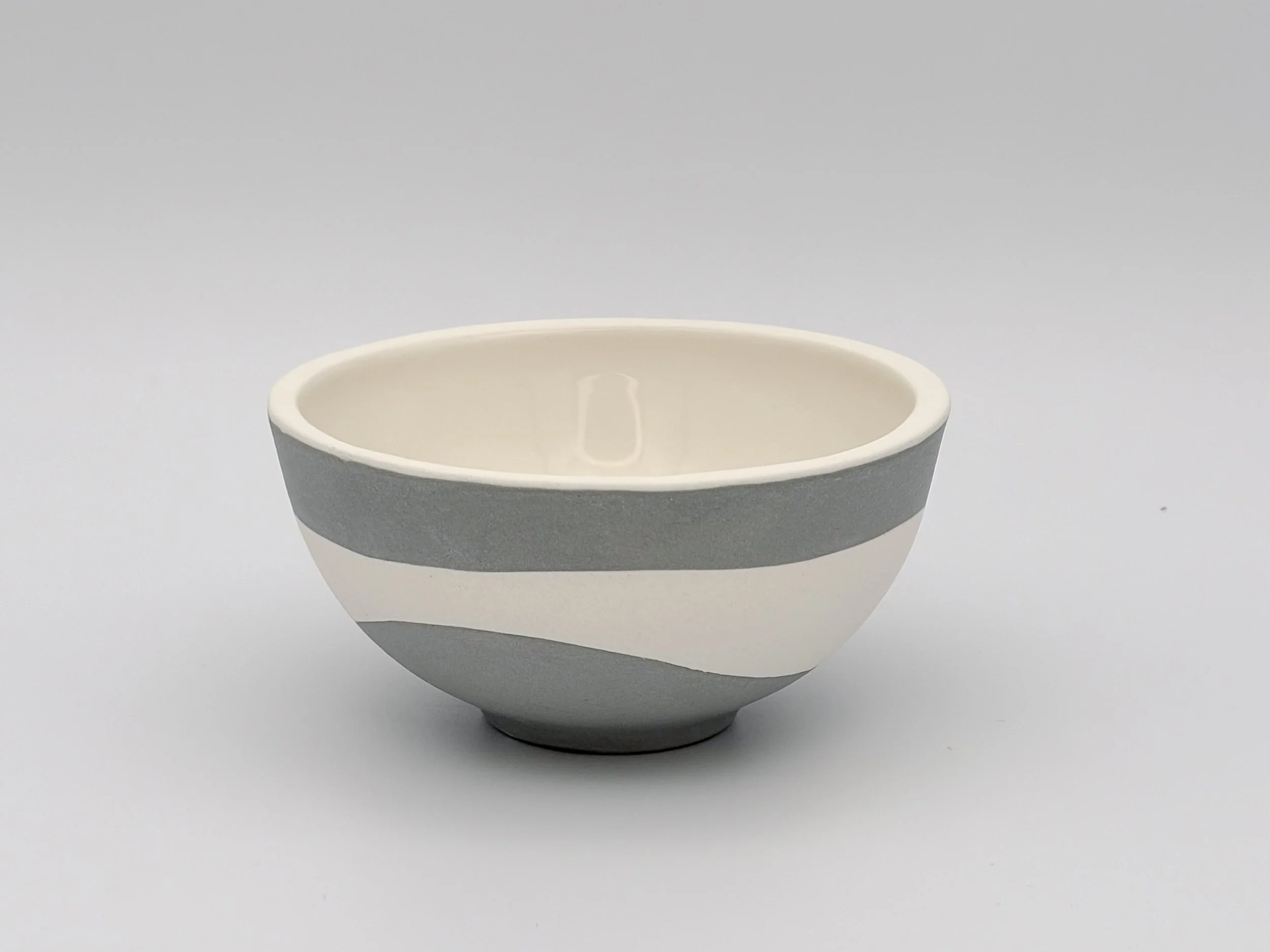 A ceramic bowl with a smooth, rounded shape and a two-tone color scheme, featuring a darker gray exterior and a light cream interior, set against a plain white background.