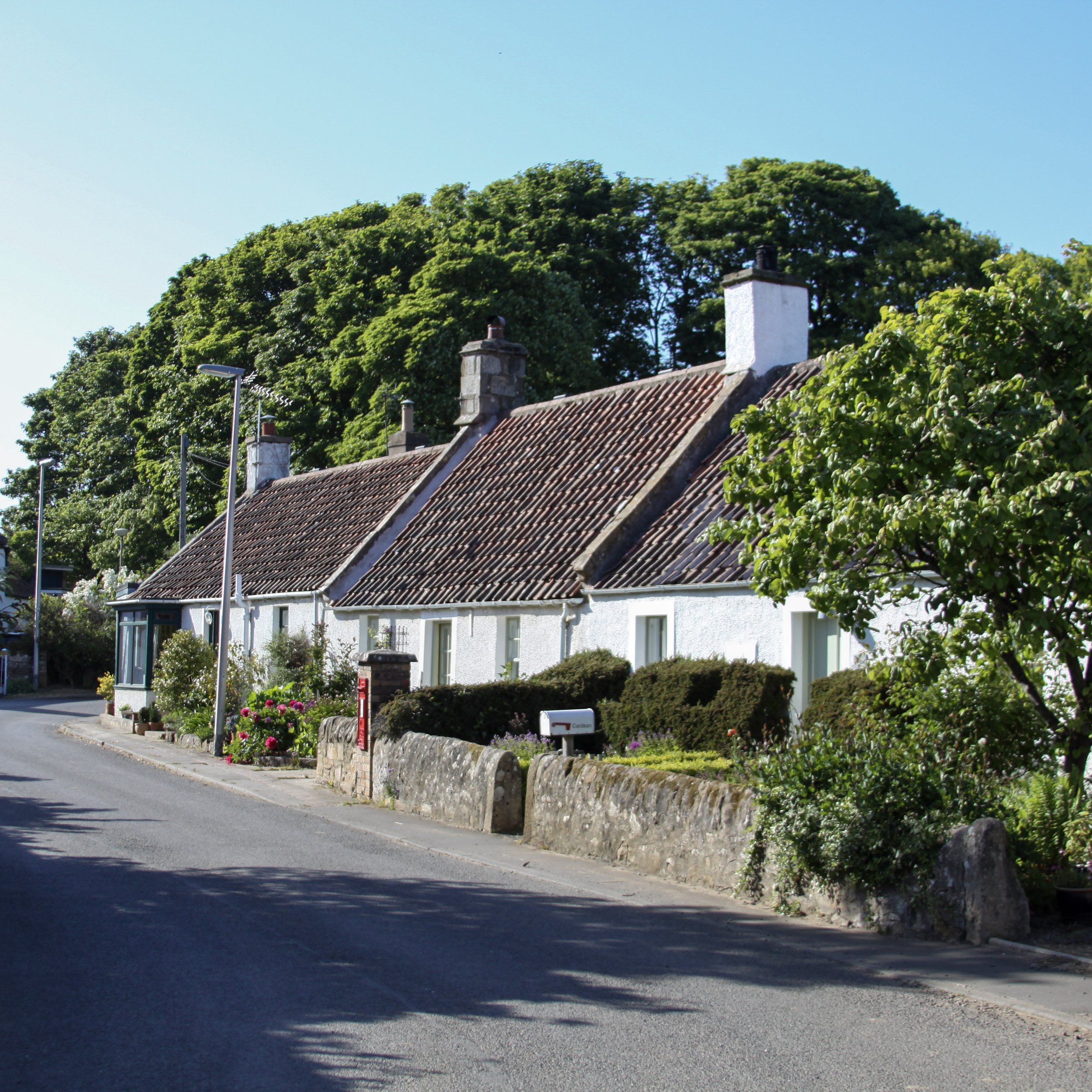 A row of white cottages with tiled roofs along a quiet street, surrounded by greenery and trees under a clear blue sky.