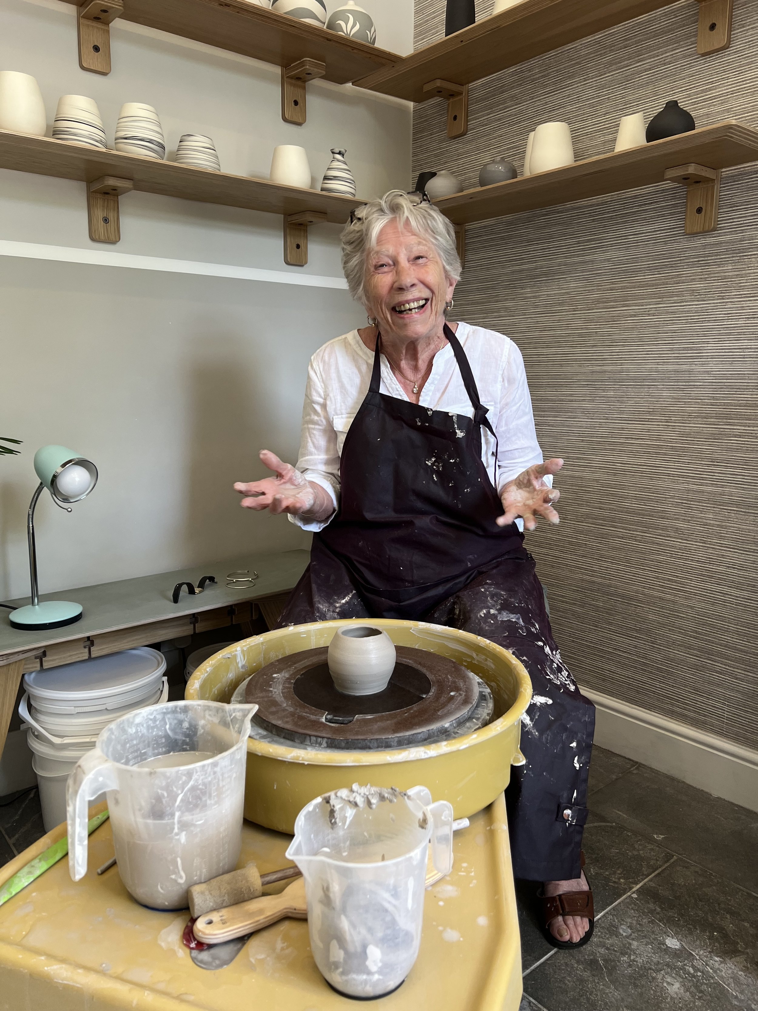A woman in a black apron is sitting at a pottery wheel, smiling and showing her hands covered in clay. There are pottery tools, pitchers with water, and clay on her lap and table, indicating she is in the process of making pottery.