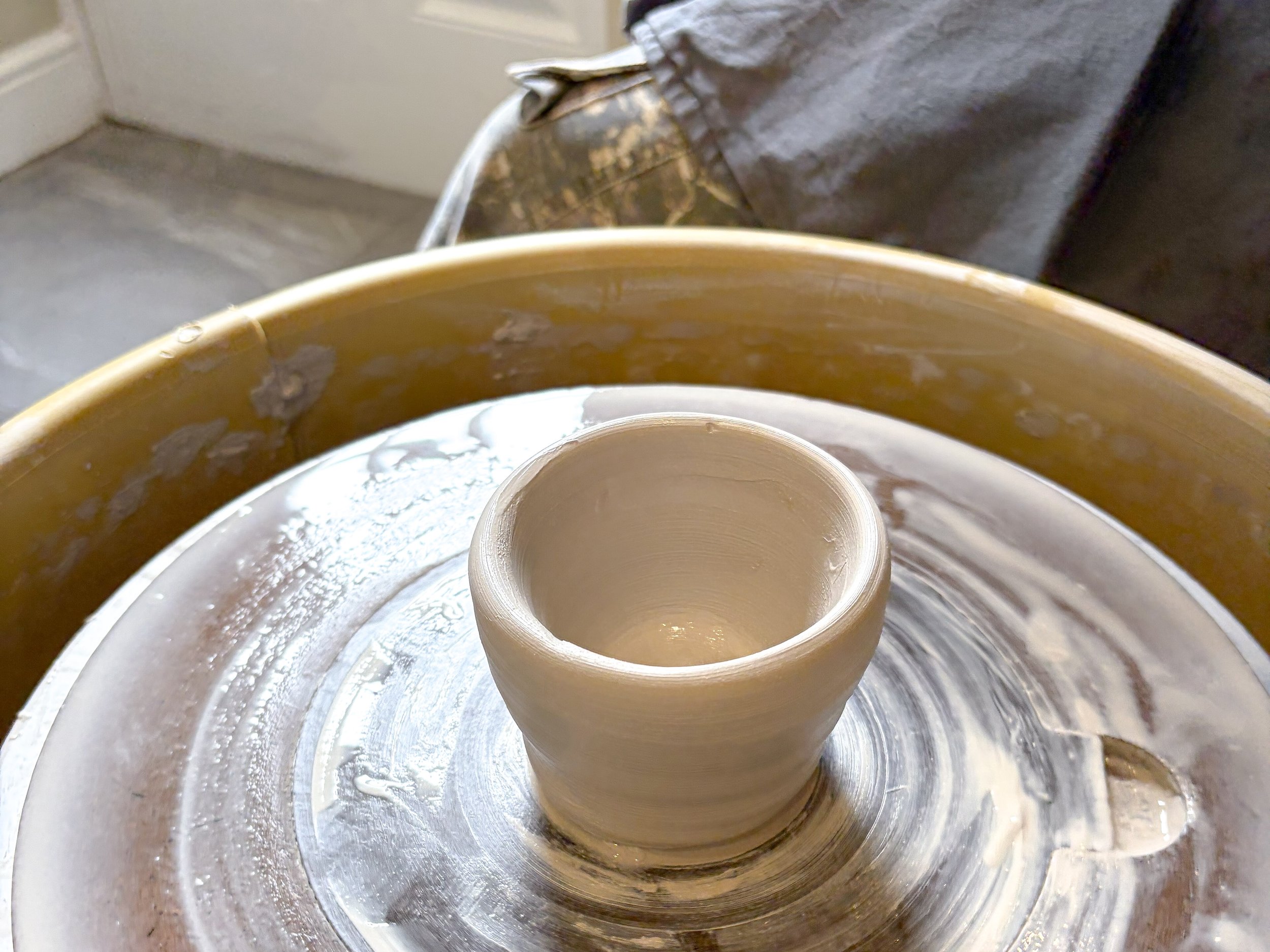 A pottery wheel with a partially formed clay mug in the center, surrounded by clay splatters and a gray cloth in the background.
