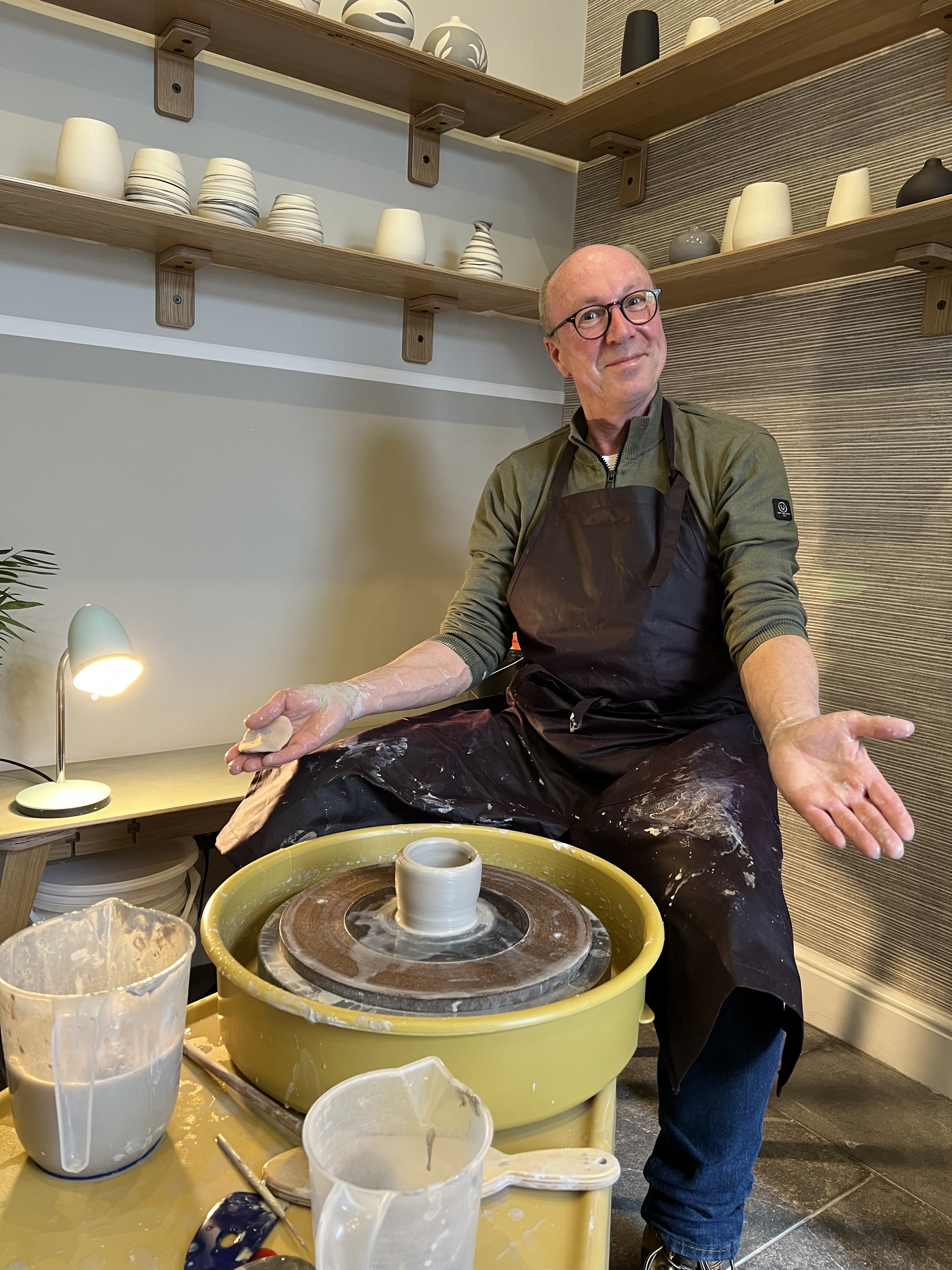 A man wearing glasses and a black apron sitting at a pottery wheel, shaping a clay mug, with pottery supplies surrounding him in a ceramic studio.