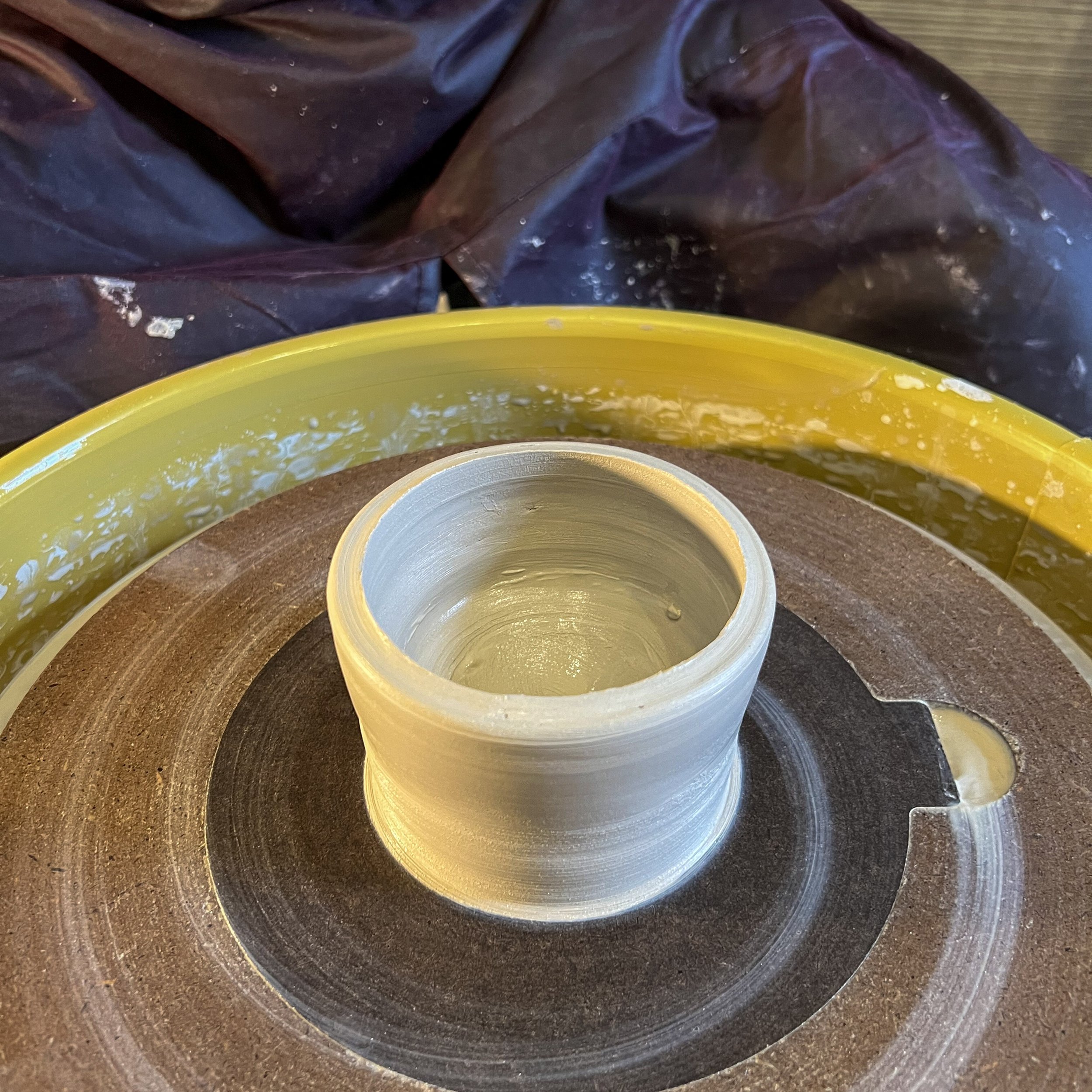 A close-up of a pottery wheel with a cylindrical ceramic piece in progress, surrounded by a yellow splash guard and a black protective cloth in the background.