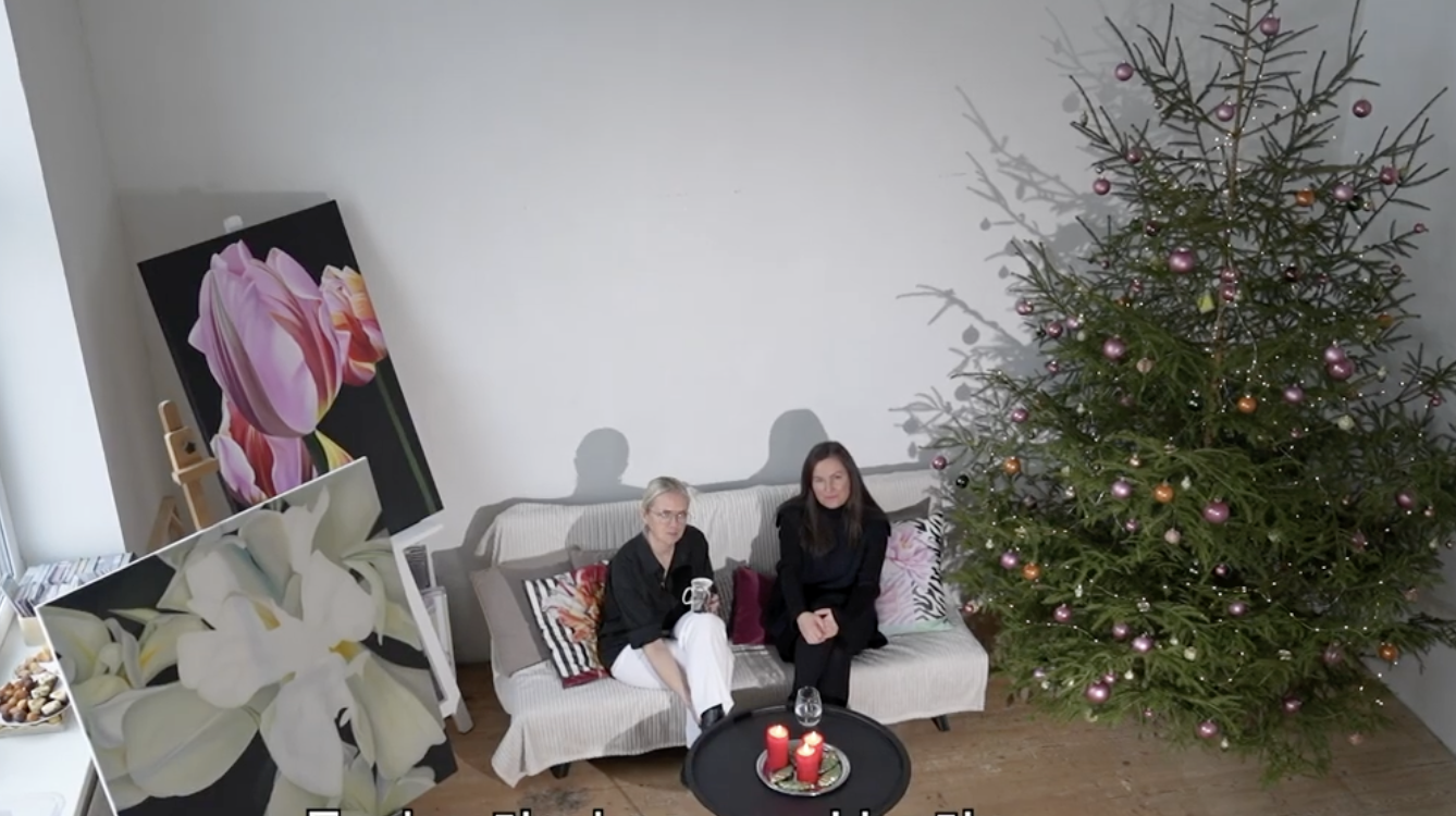 Two women sitting on a sofa next to a decorated Christmas tree in a living room. There are paintings of flowers leaning against the wall.