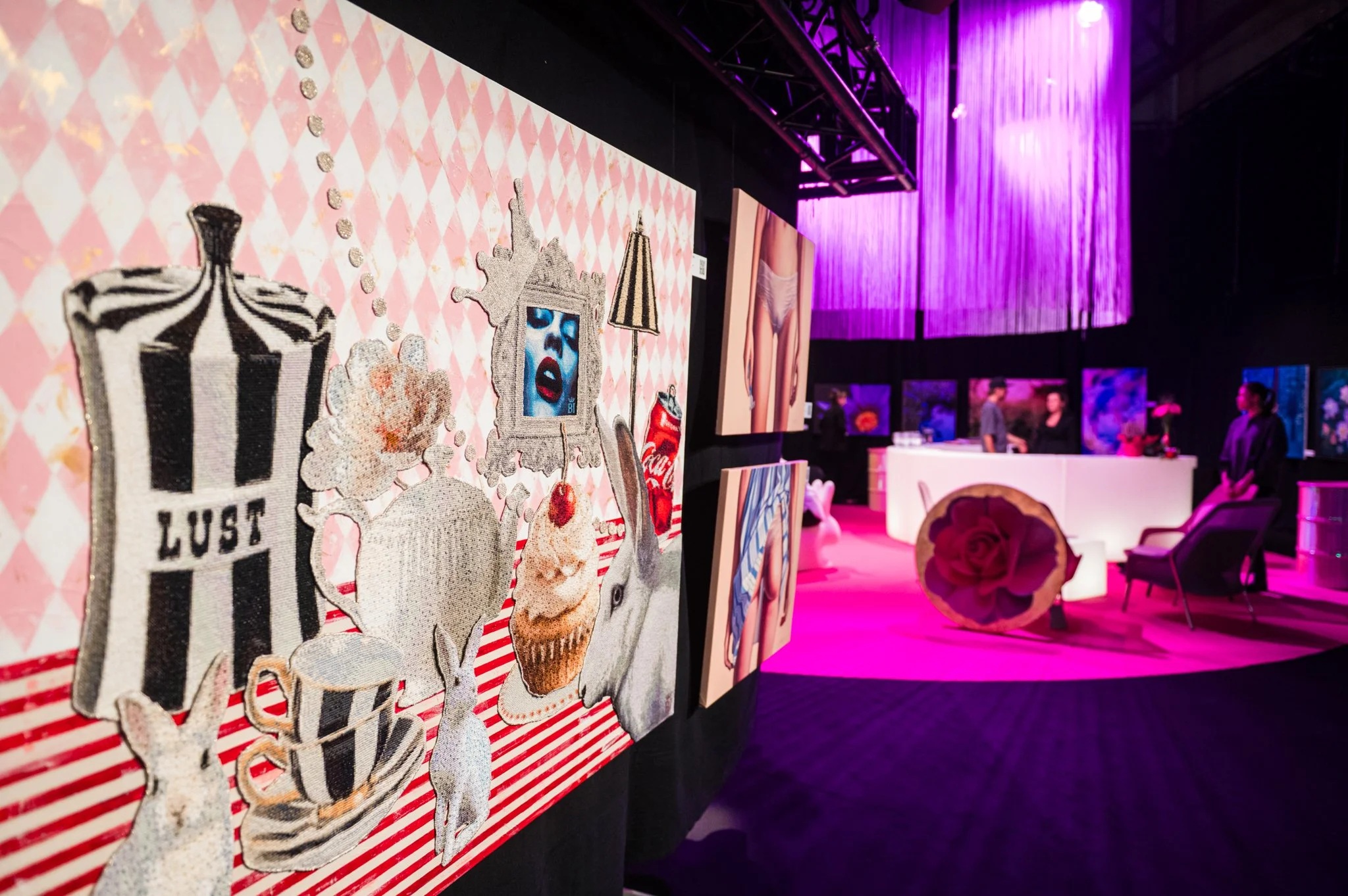 Art exhibit with rhinestone-encrusted embroidery of various pop art and whimsical images on a pink diamond-patterned background, displayed on a wall in a gallery with dark walls, purple lighting, and artwork in the background.