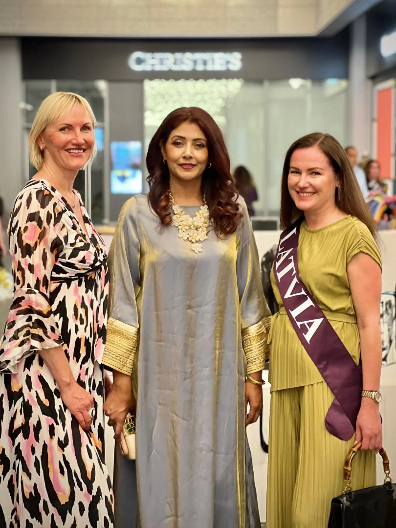 Three women standing together in an indoor setting, one wearing a leopard print dress, another in a traditional outfit with gold accents and statement necklace, and a third wearing a green dress with a sash that reads 'VIVAT'.