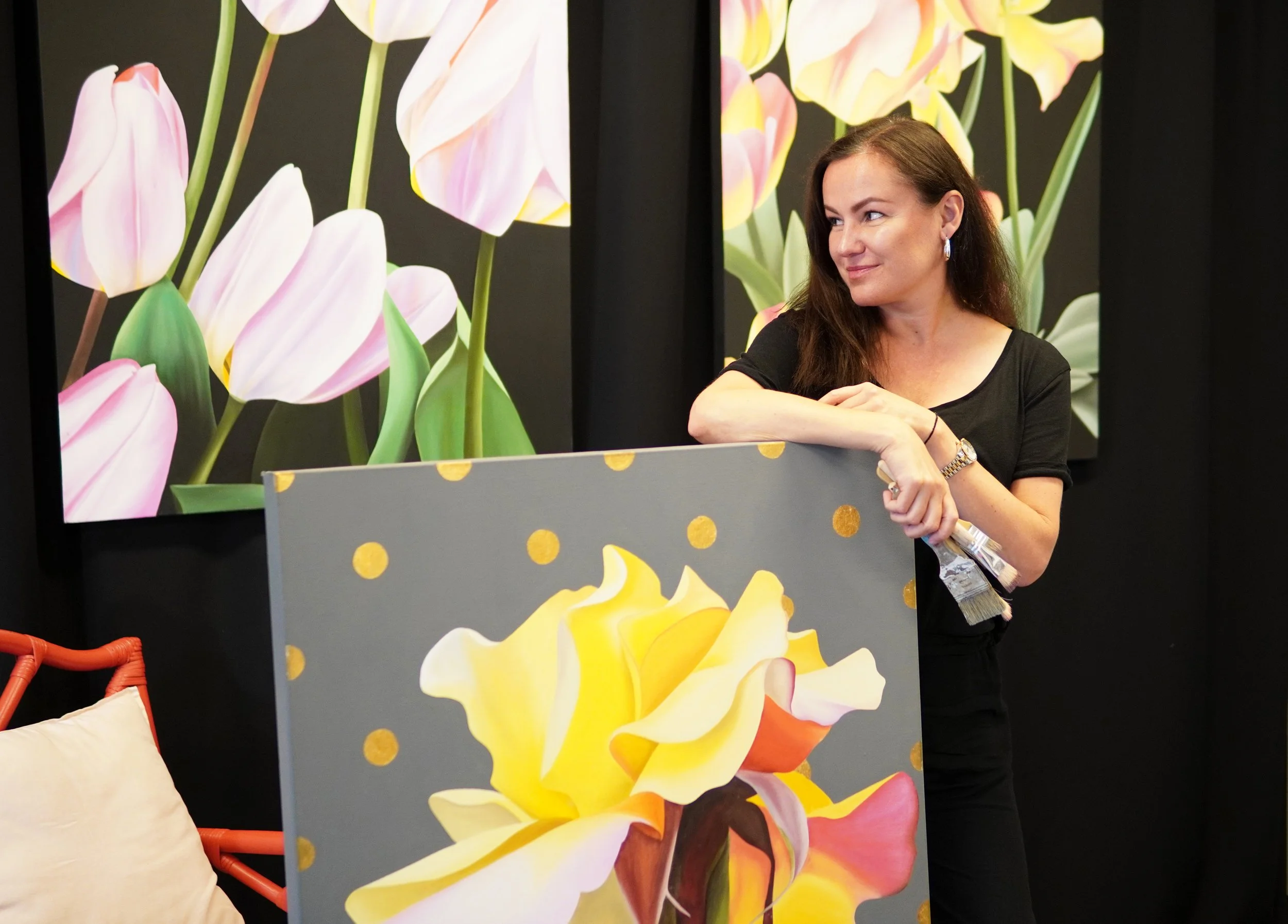 A woman with long dark hair and a black shirt standing beside a painting of a yellow flower with pink and orange accents, in an art gallery with floral artwork on the walls.