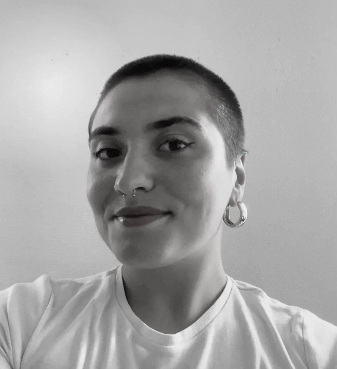 Portrait d'une personne avec une coupe de cheveux courte, portant des boucles d'oreilles en forme de anneau et un piercing au septum. Photo en noir et blanc.