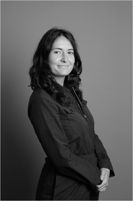 Portrait en noir et blanc d'une femme avec des cheveux bouclés, portant une tenue sombre à rayures, posant en souriant légèrement, face à un fond uni.