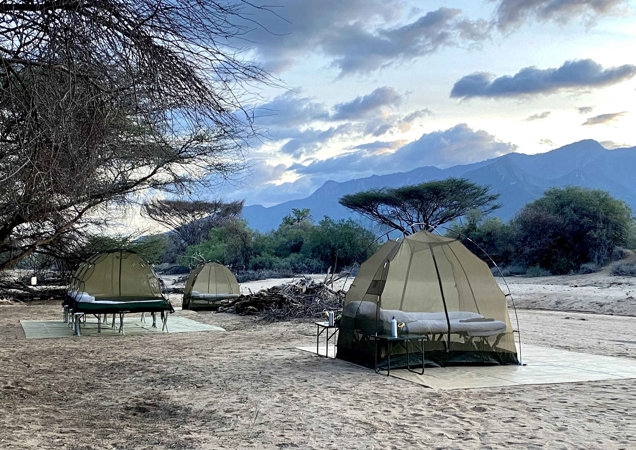 Camping tents set up on a sandy area with a backdrop of trees, mountains, and a cloudy sky at sunset.