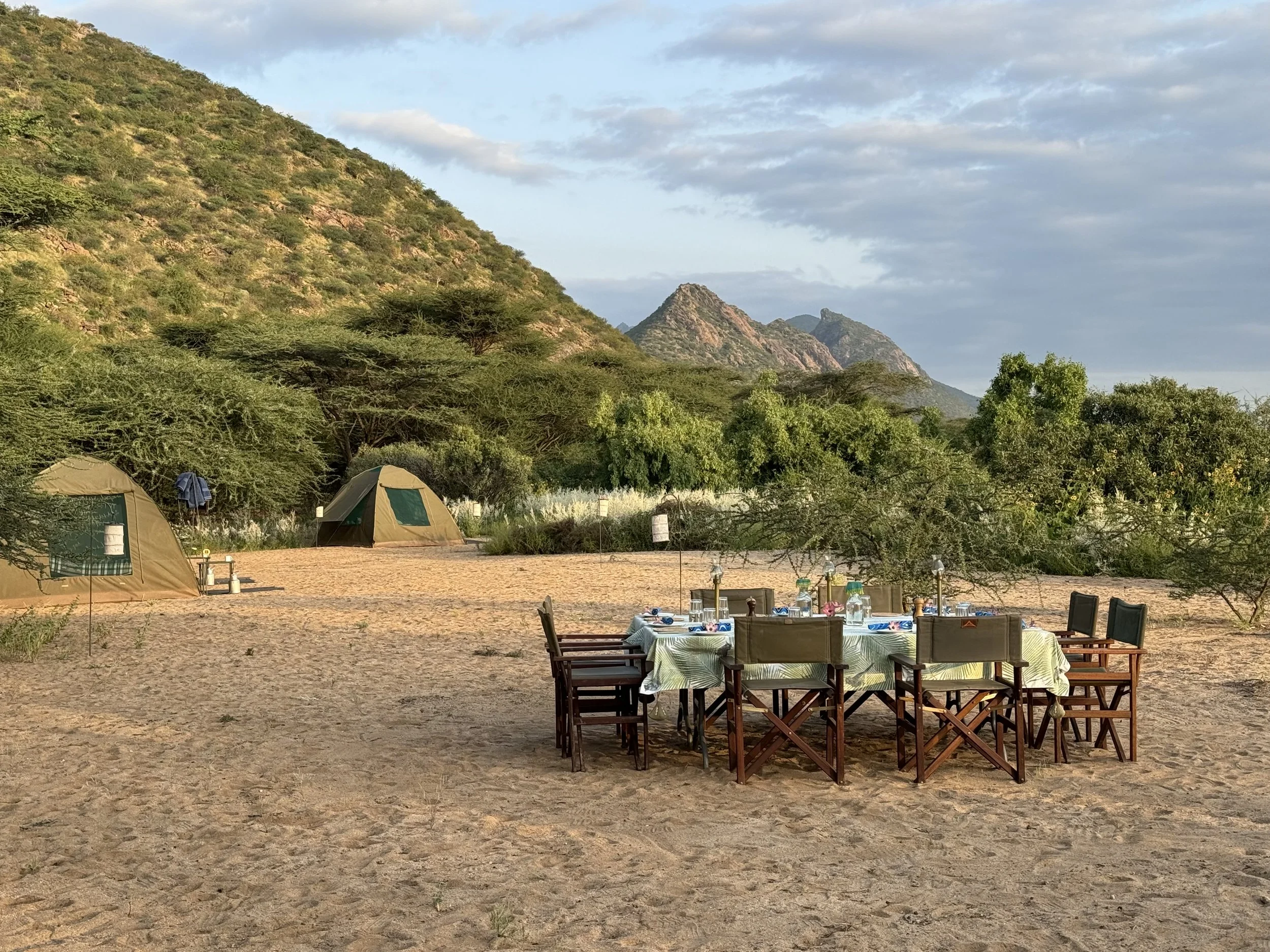 Camping scene with tents, a dining area with chairs and tables, and mountains in the background under a partly cloudy sky.