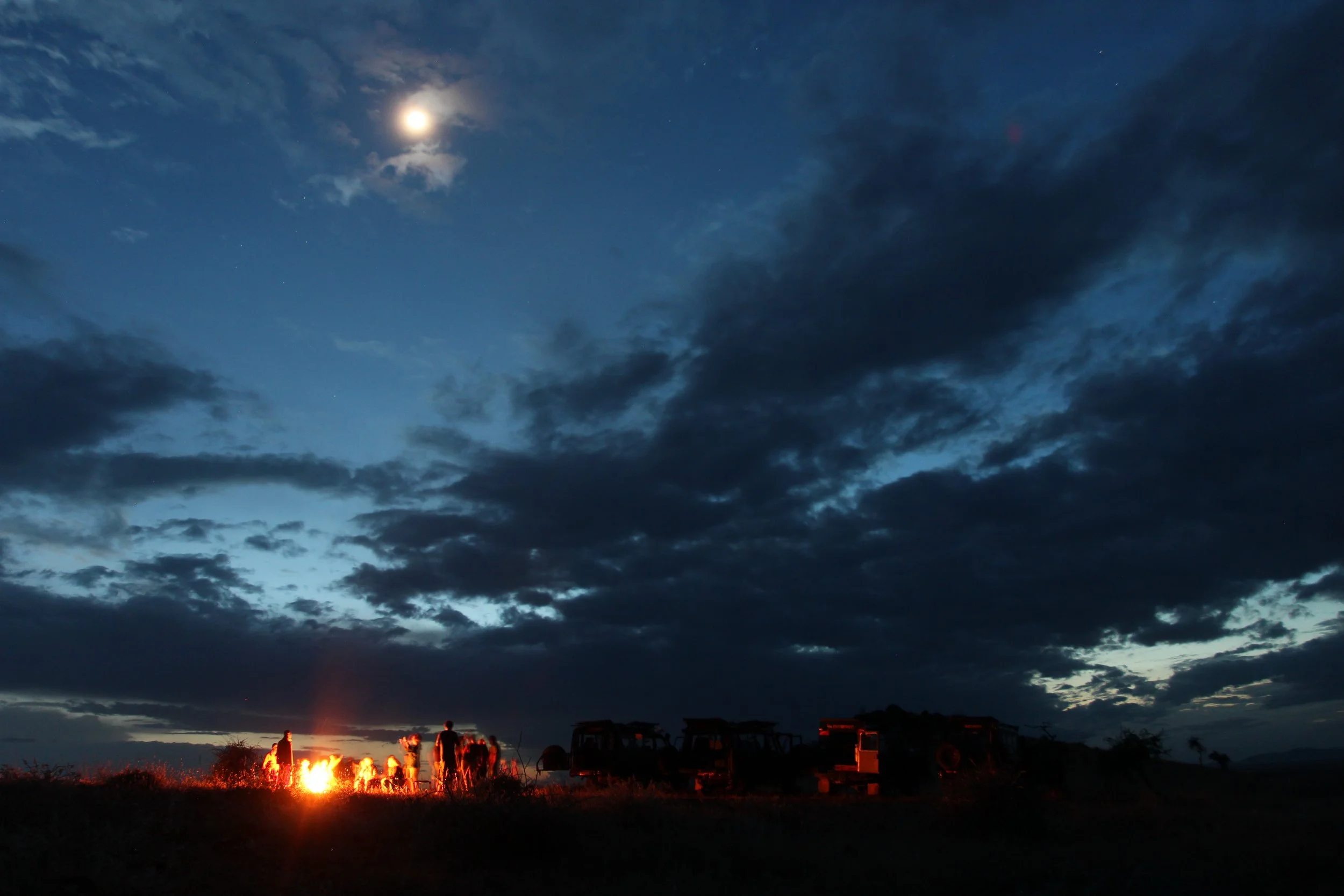 Guests gathered around a campfire enjoying a sundowner beneath the moonlit night sky during a safari in Kenya