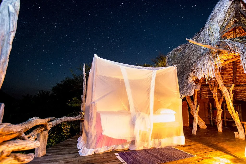 A bed with a mosquito net on a wooden deck at night, illuminated by warm lighting, surrounded by tropical trees and thatched-roof structures under a starry sky.