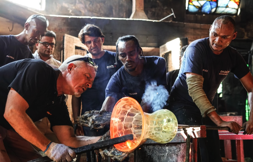Group of six men working together on glassblowing inside a workshop with stained glass window in background.