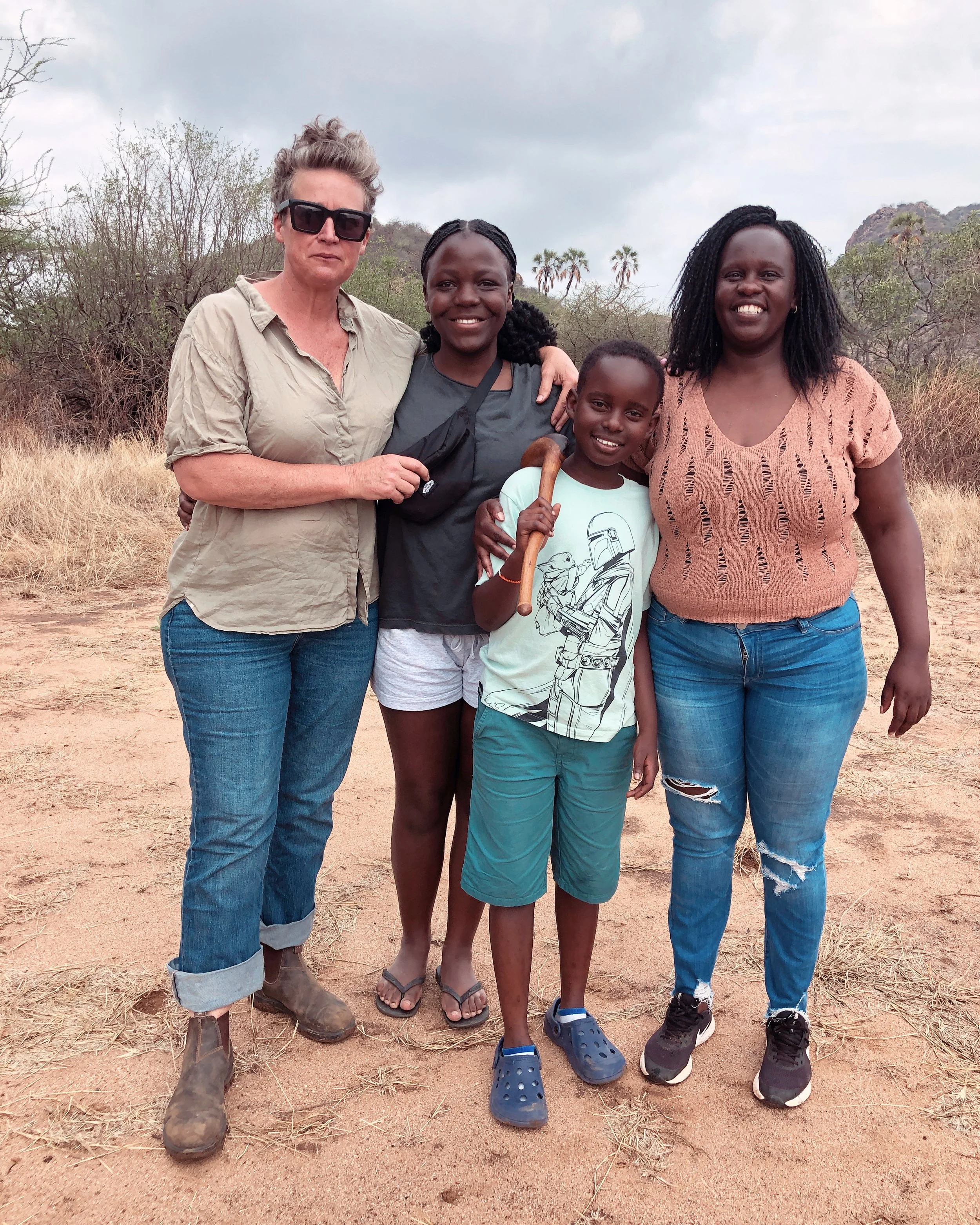 Four women of different ethnicities standing close together outdoors in a dry, grassy area with bushes and trees, smiling at the camera.