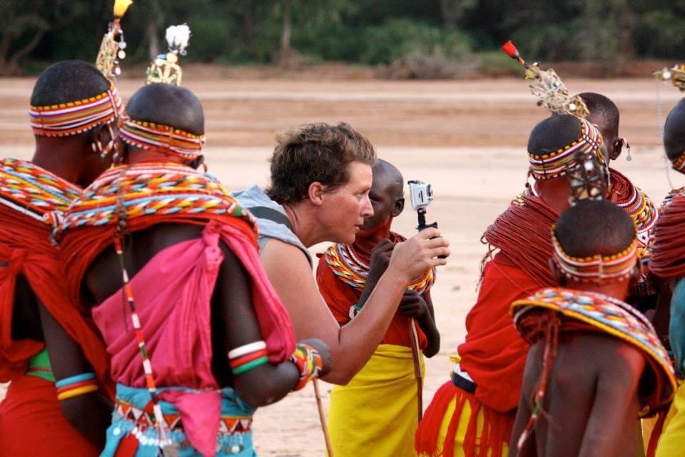 A woman taking a photograph with a small camera of a group of traditional Maasai tribesmen dressed in colorful clothing and jewelry.
