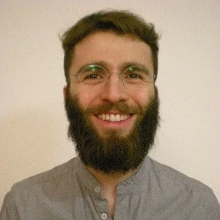 A smiling man with glasses and a beard wearing a gray shirt in front of a plain wall.