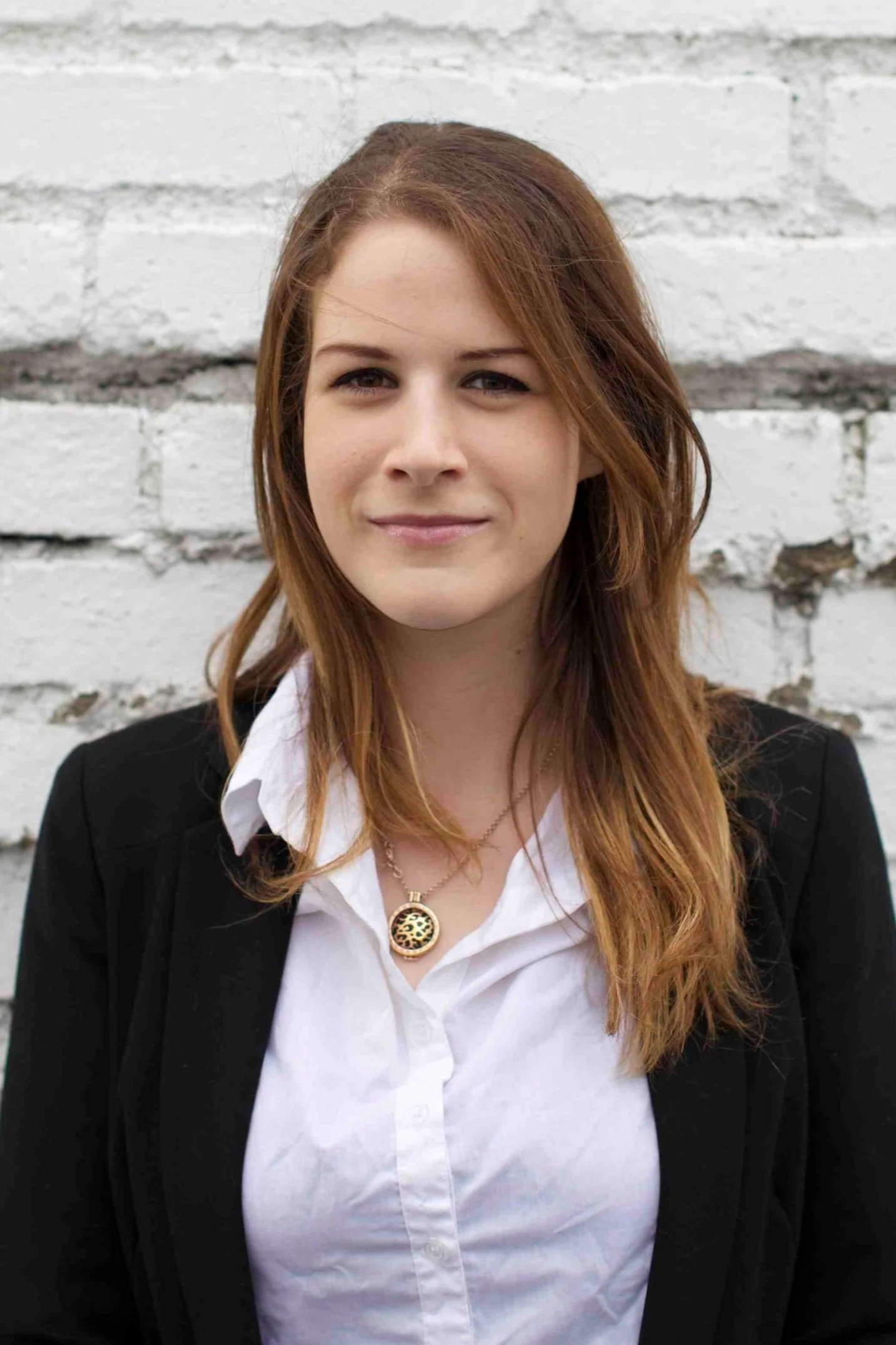 A woman with long red hair and fair skin, wearing a black blazer over a white shirt with an open collar, standing in front of a white brick wall.