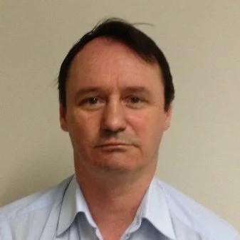 A man with short dark hair wearing a light-colored button-up shirt, standing against a plain beige wall.