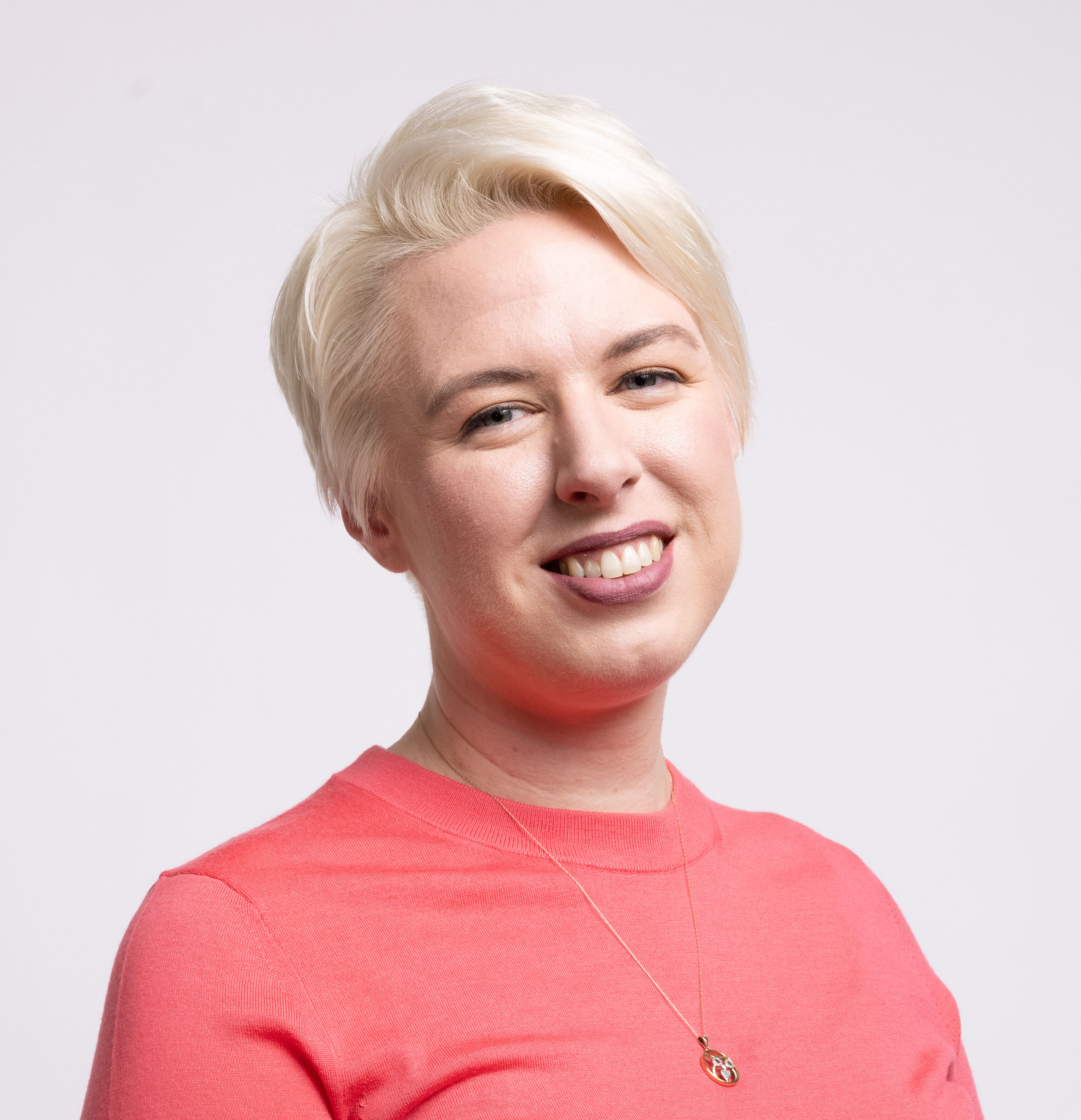 A woman with short, platinum blonde hair smiling, wearing a salmon-colored top and a gold necklace.