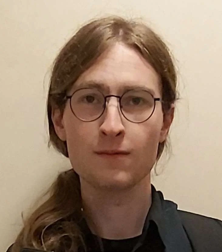 A young person with long brown hair, glasses, and wearing a black shirt, standing against a beige wall.