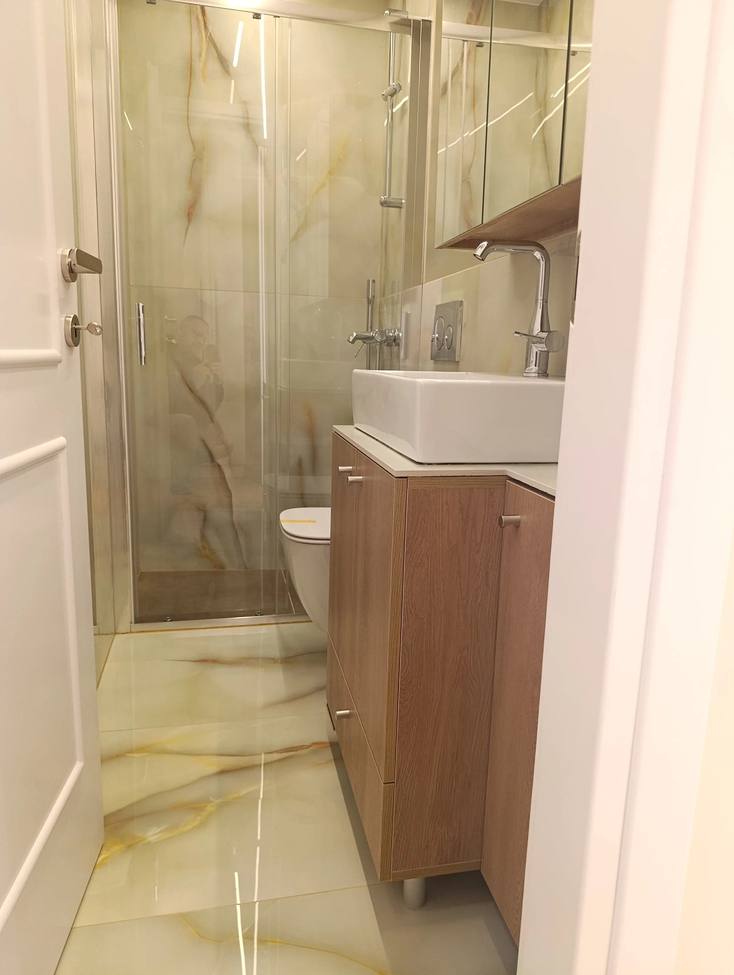 A compact bathroom with a glass shower enclosure, a wall-mounted toilet, a white rectangular vessel sink on a wooden vanity, a mirror, and marble-like tiled walls and floor.