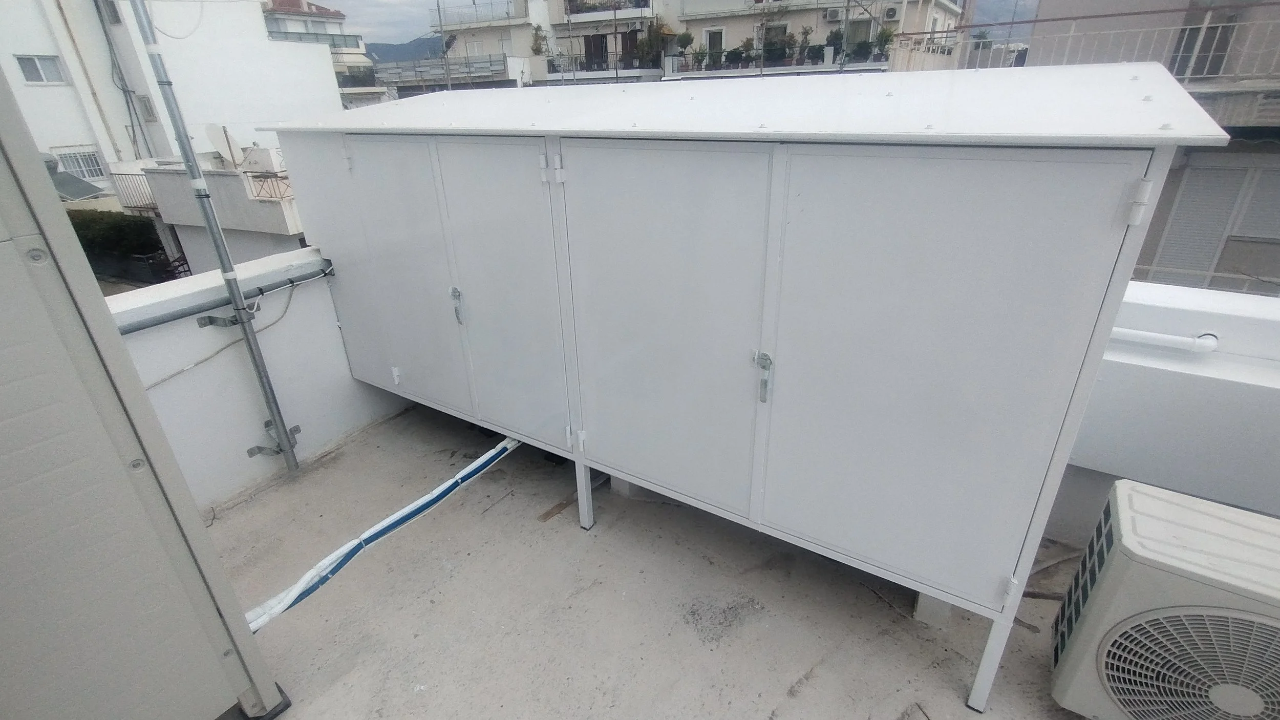 Outdoor rooftop area with a large white electrical or utility cabinet, pipes, and an air conditioning unit on the ground.
