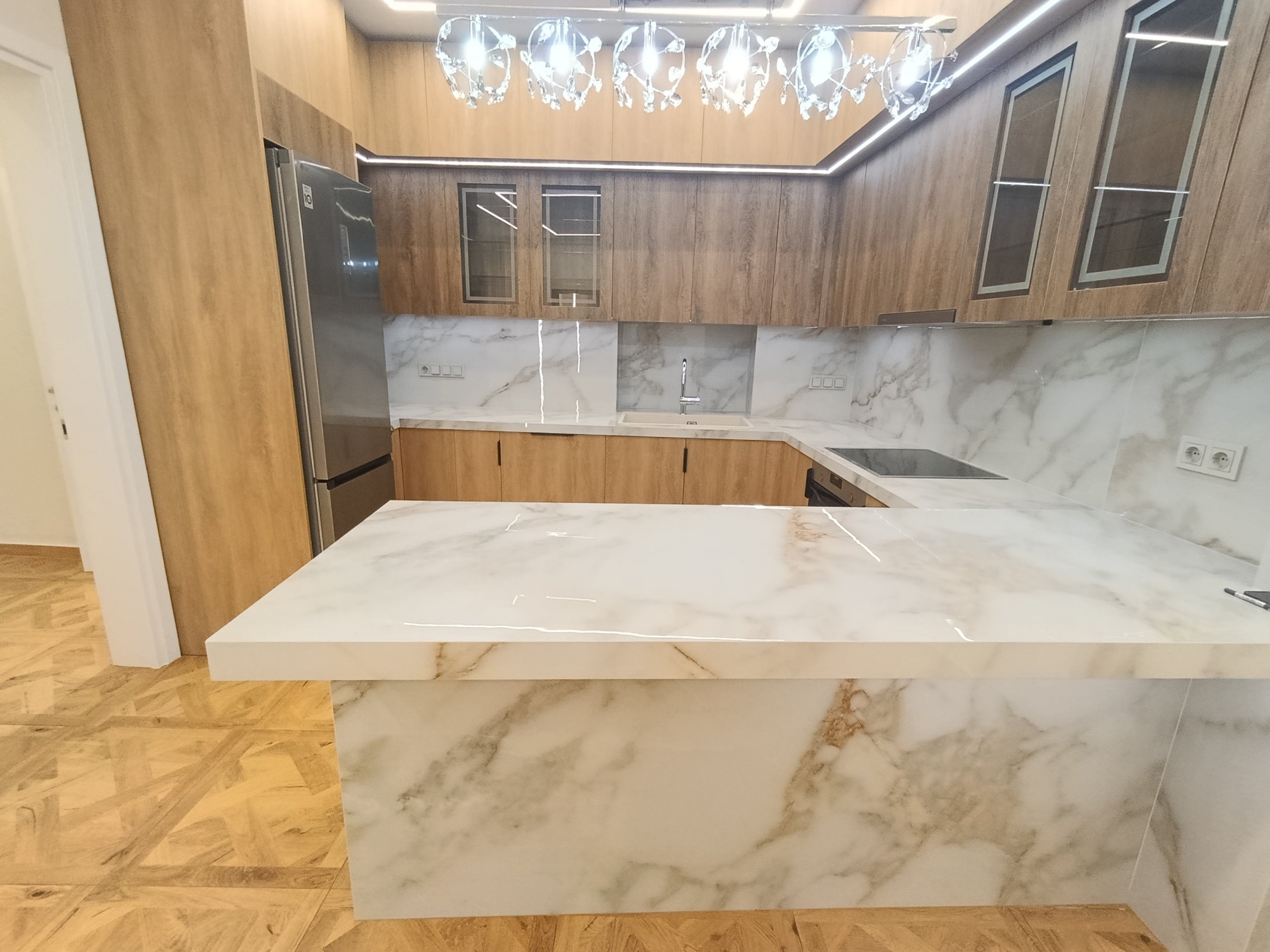 Modern kitchen with wood cabinets, marble countertops, and a marble island. Stainless steel appliances and a chandelier above.