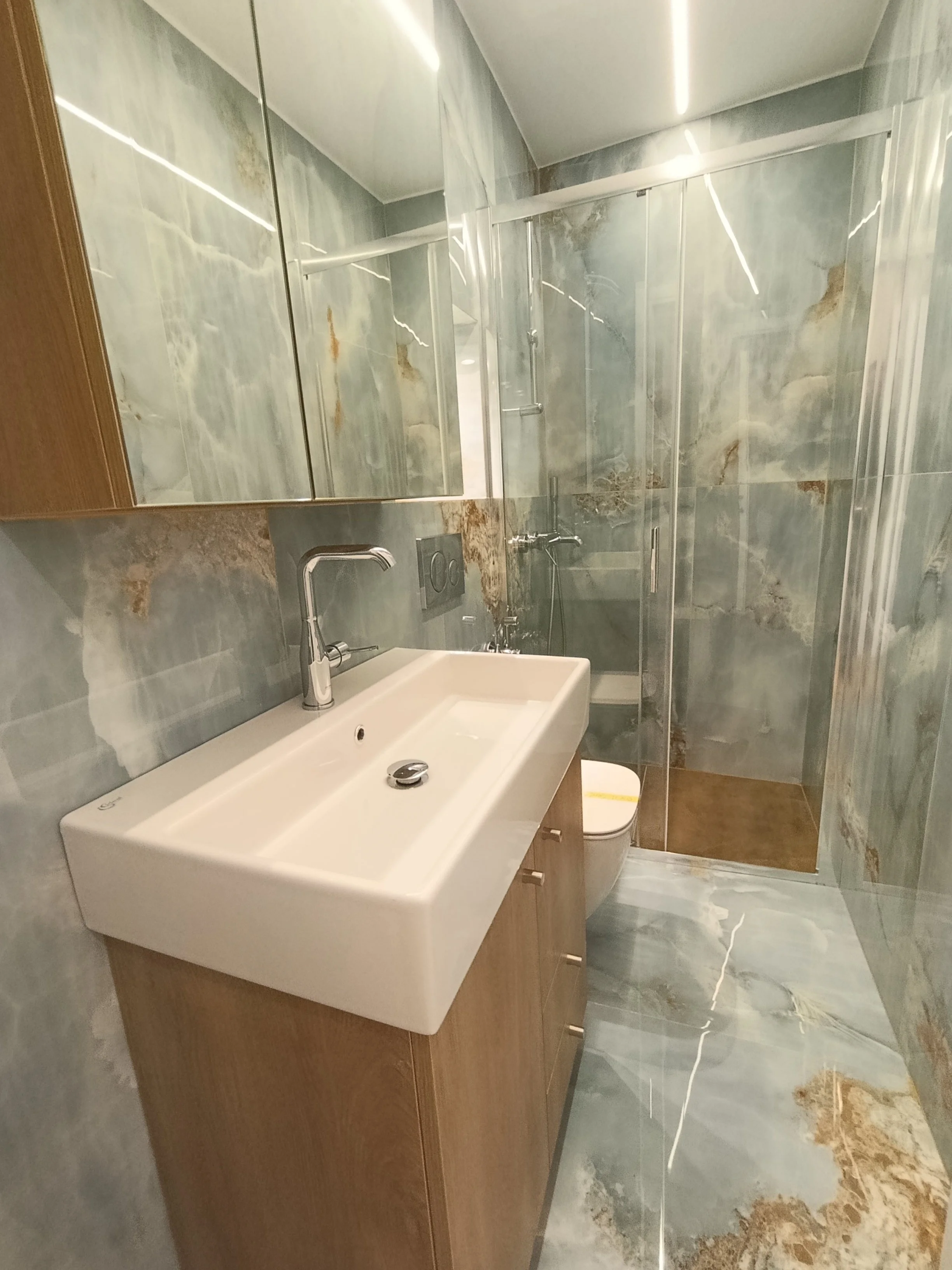 Modern bathroom with marble walls and floors, a white sink with wooden cabinet, a mirror, a toilet, and a glass shower enclosure.