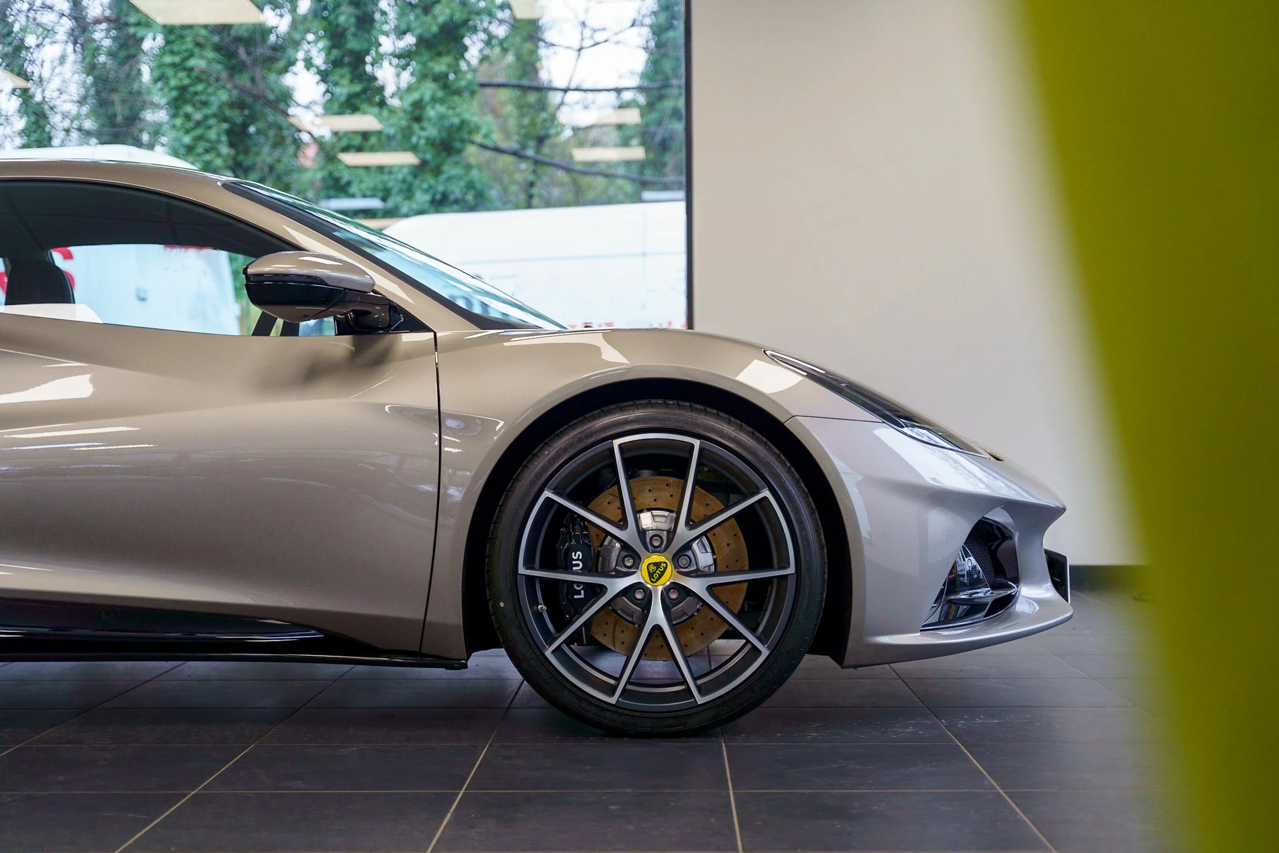 Close-up of a silver sports car displayed indoors, showing the front wheel, part of the side, and a large brake caliper, with a window and trees in the background.