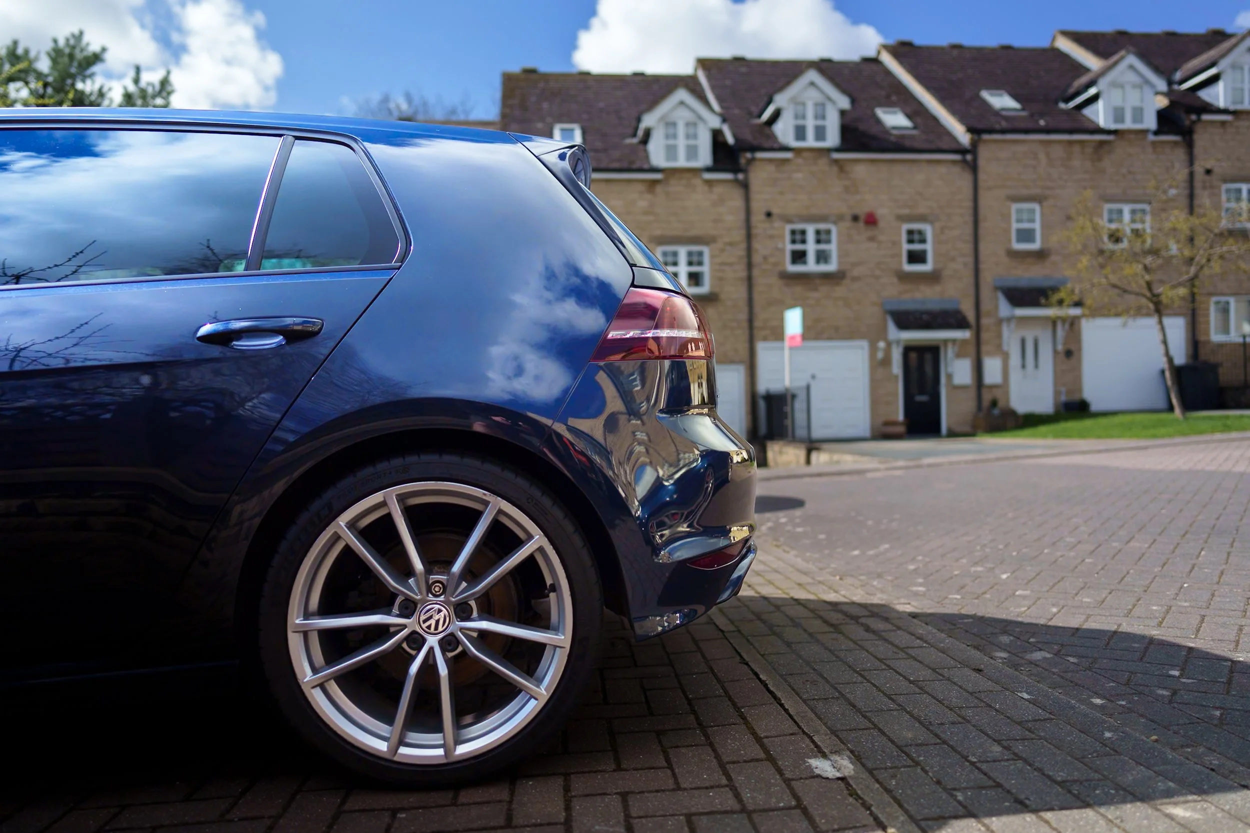 Rear view of a dark blue Volkswagen car parked on a brick driveway in front of modern townhouses on a sunny day.
