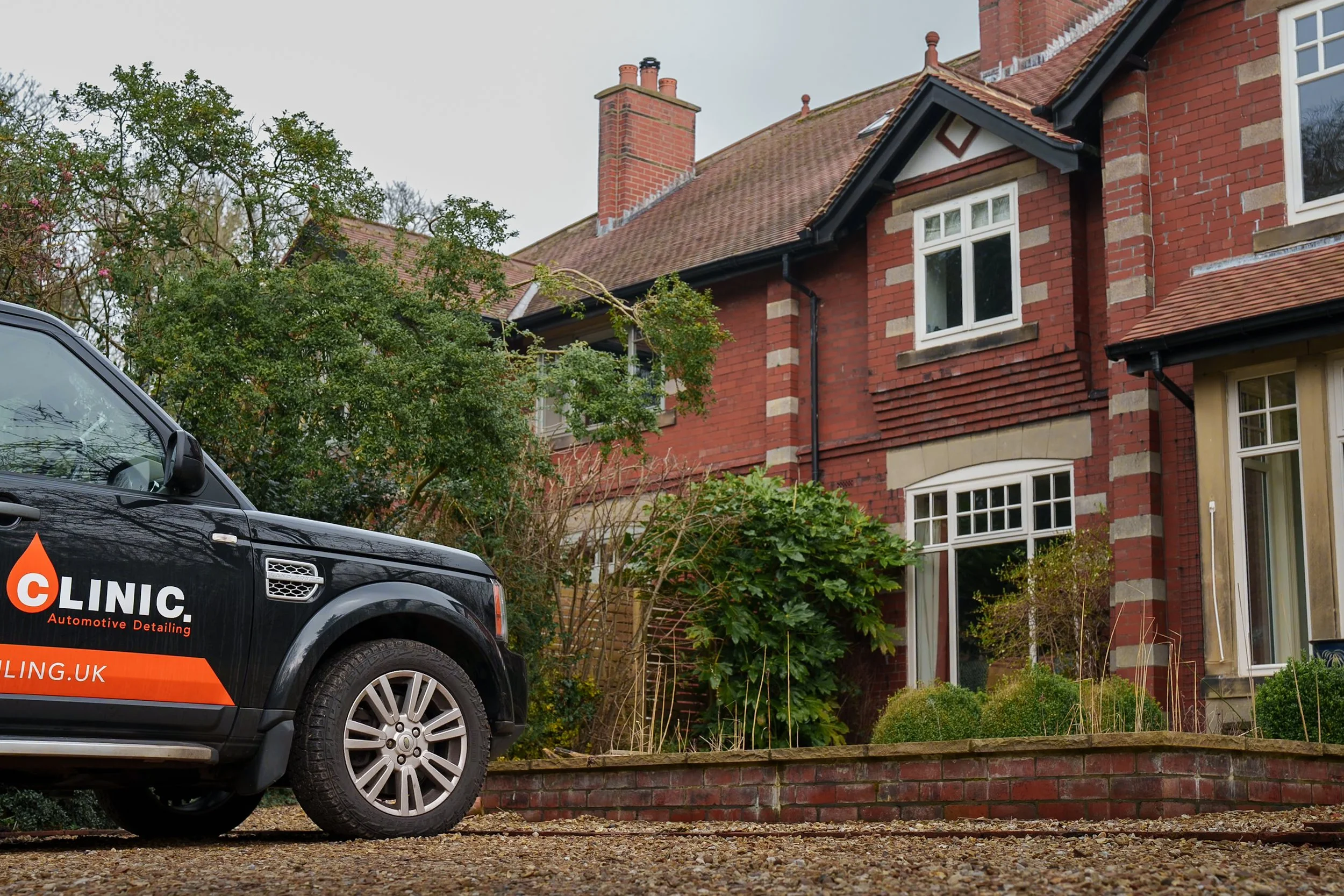 A black vehicle parked in front of a red-brick house with white window frames and a garden with bushes and plants.