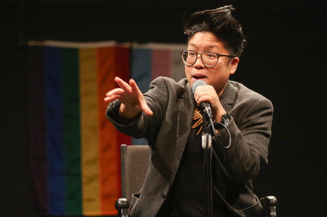 A brown, round queer performs at a microphone with transgender and rainbow flags in the background. They wear a gray blazer and glasses.