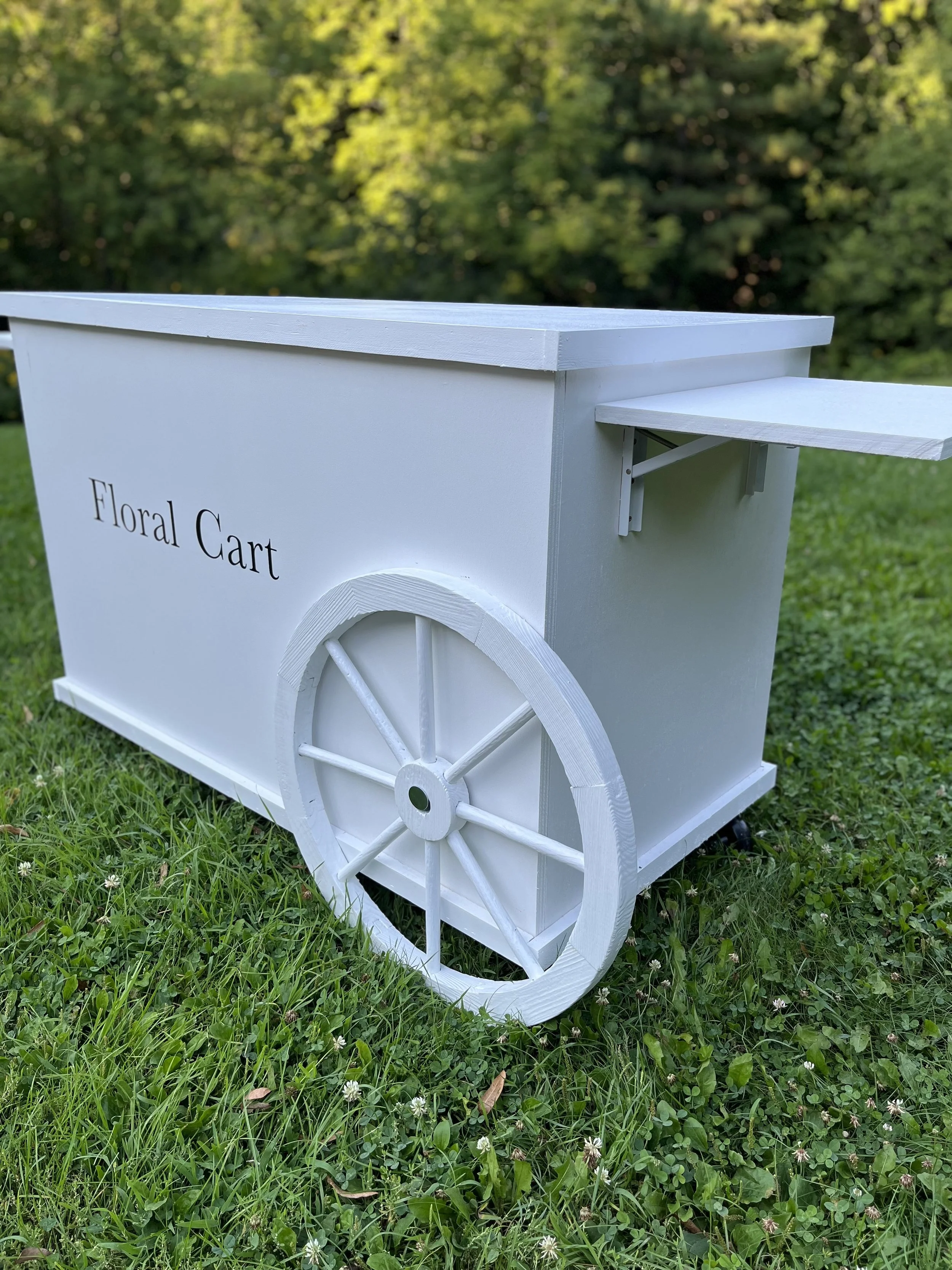 White wooden floral cart with large wheel, decorated for an event, labeled 'Floral Cart', outdoors on a grassy area with trees in the background.