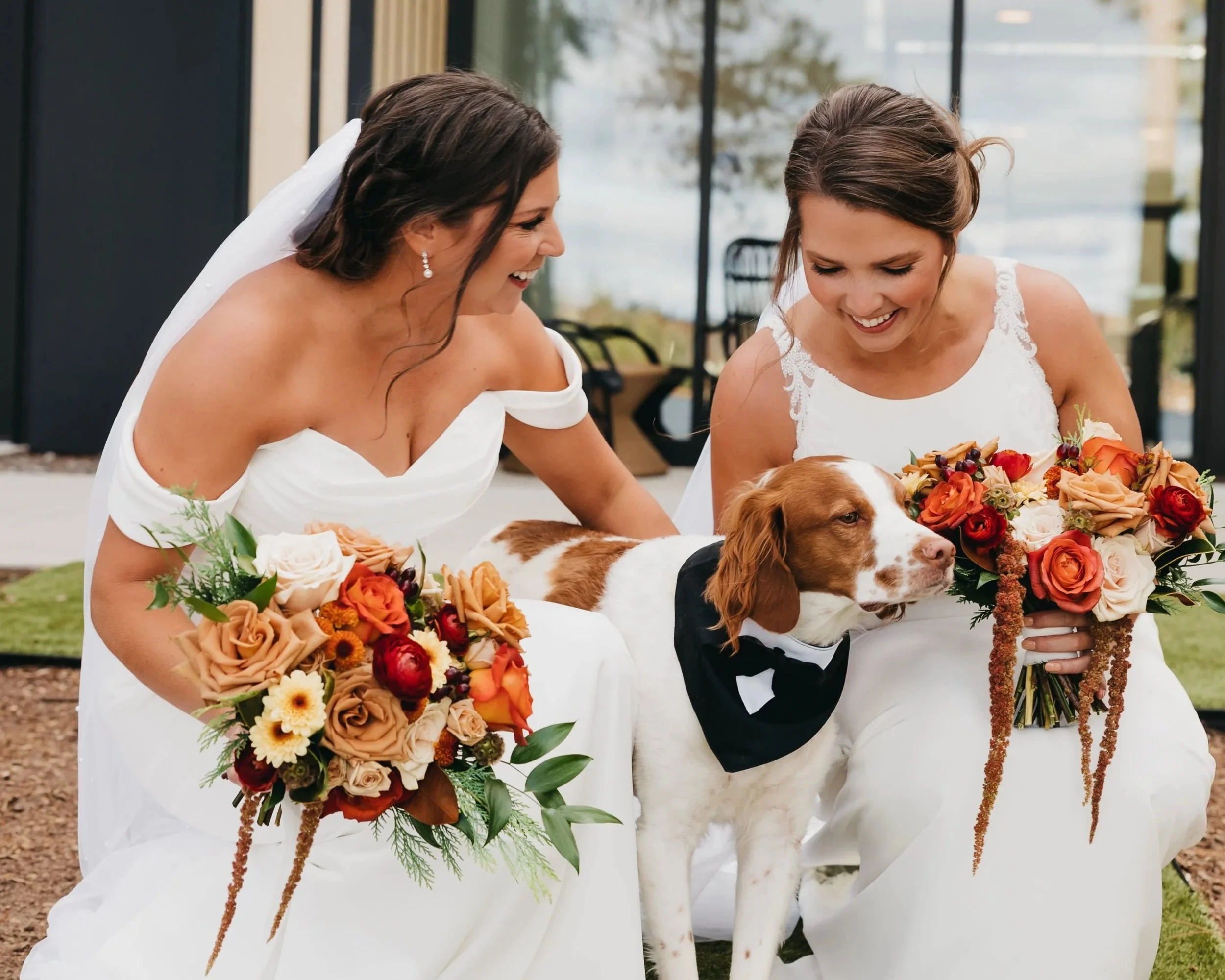 Two brides crouching with their bouquets and their dog at an outdoor Nisswa, Minnesota wedding.